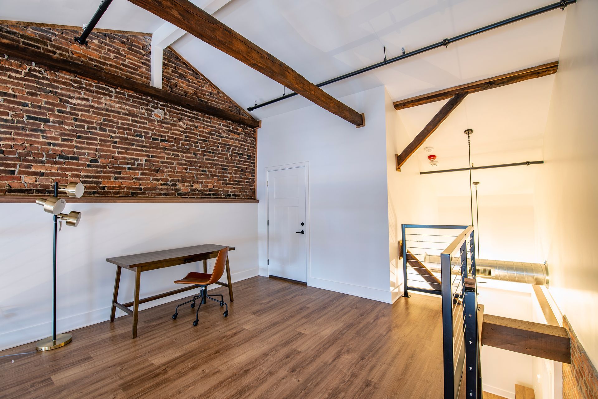 Empty attic room with exposed brick wall, wooden beams, and a small table by a bright window.