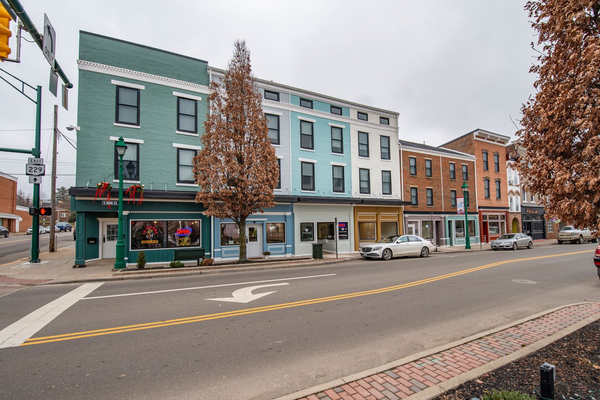 Small-town street with colorful storefronts, parked cars, and autumn trees under an overcast sky.