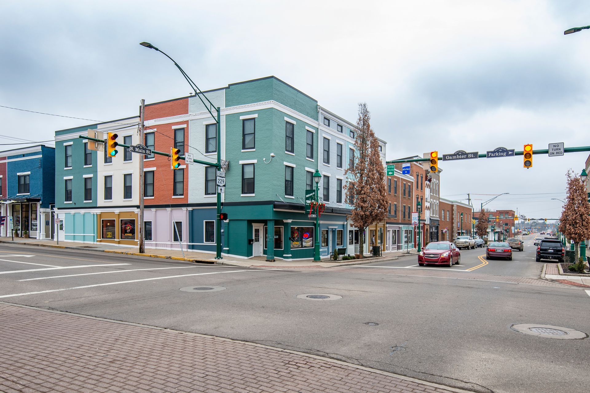Colorful corner buildings at a city intersection with cars, traffic lights, and a cloudy sky.