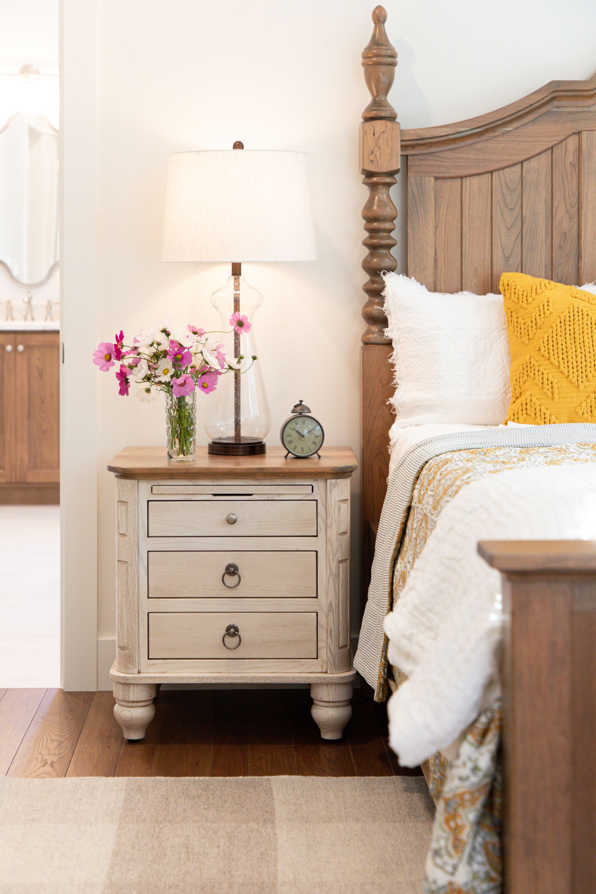 Cozy bedroom with a carved bed, beige nightstand, lamp, and pink flowers by the bedside.