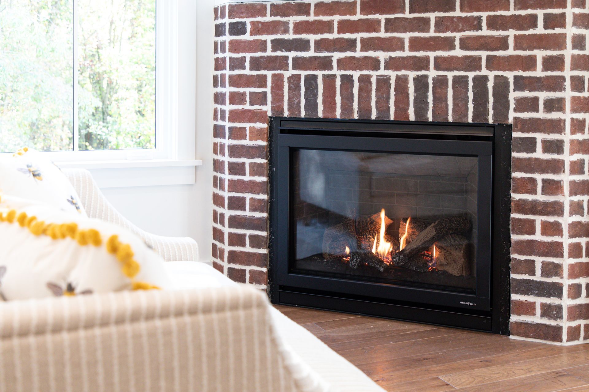 Cozy living room with lit fireplace, brick surround, and striped armchair by a bright window.