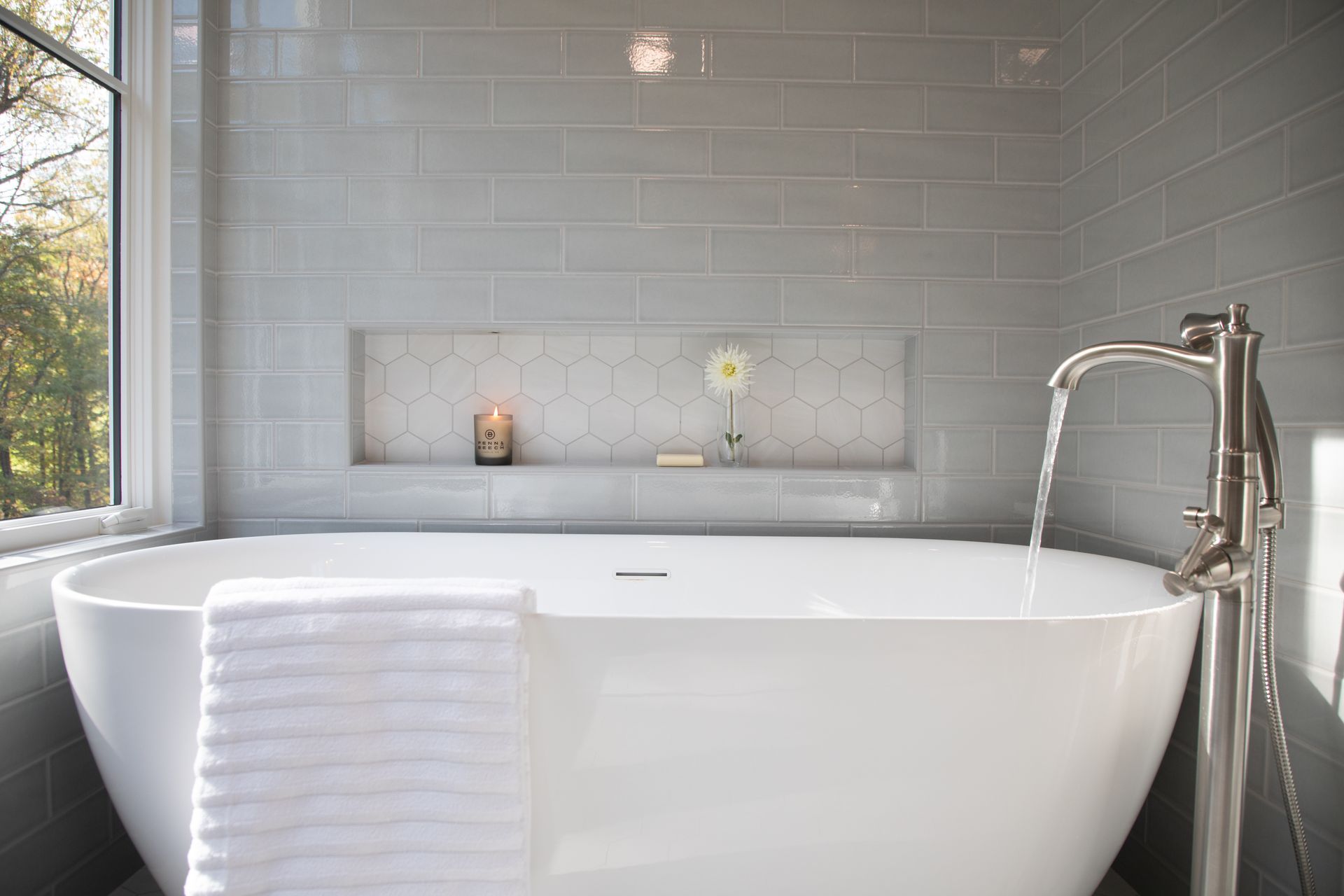 Modern white bathroom with freestanding tub, chrome faucet, window, and built-in shelf with candles and flowers.