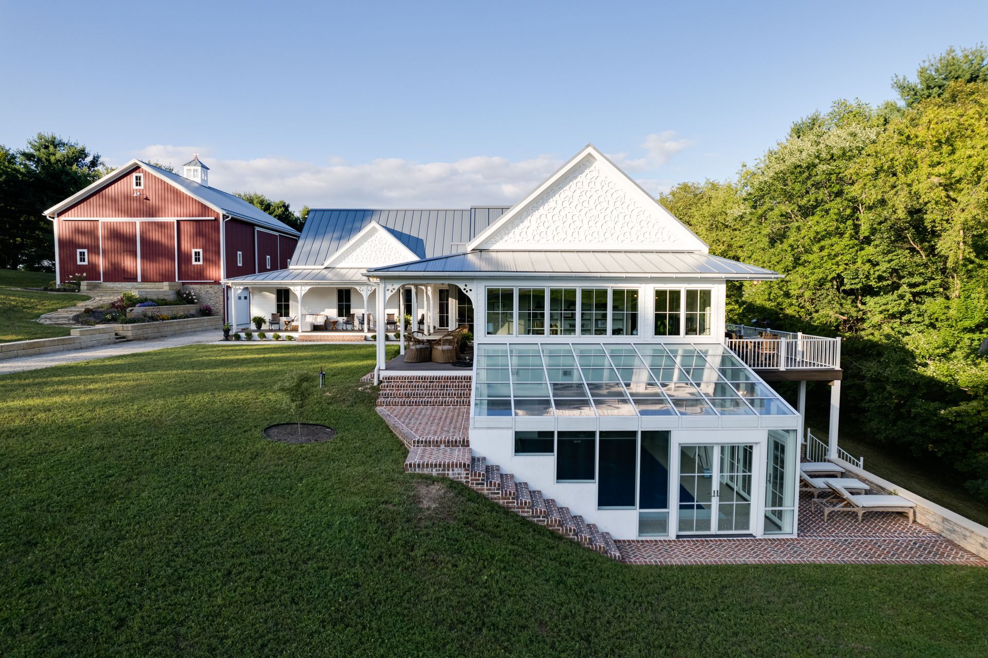 White house with large glass sunroom beside a red barn on a grassy hillside.