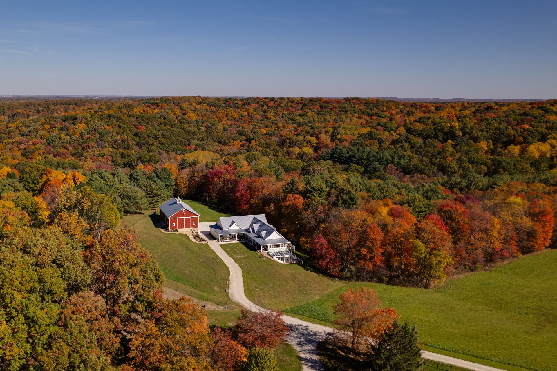 Aerial view of a secluded custom farmhouse and winding driveway surrounded by colorful autumn forest and fields.