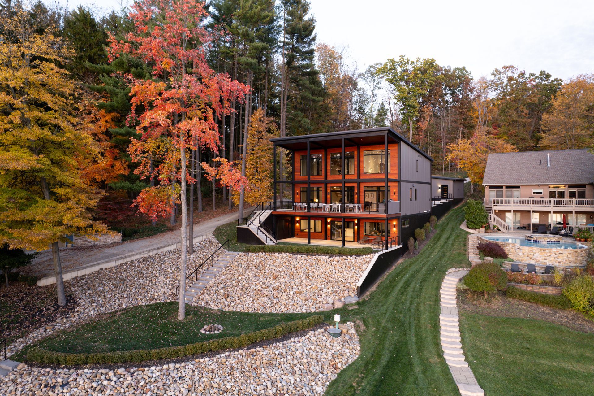 Modern custom house with glass walls beside a stone-lined riverbank in a wooded autumn landscape.