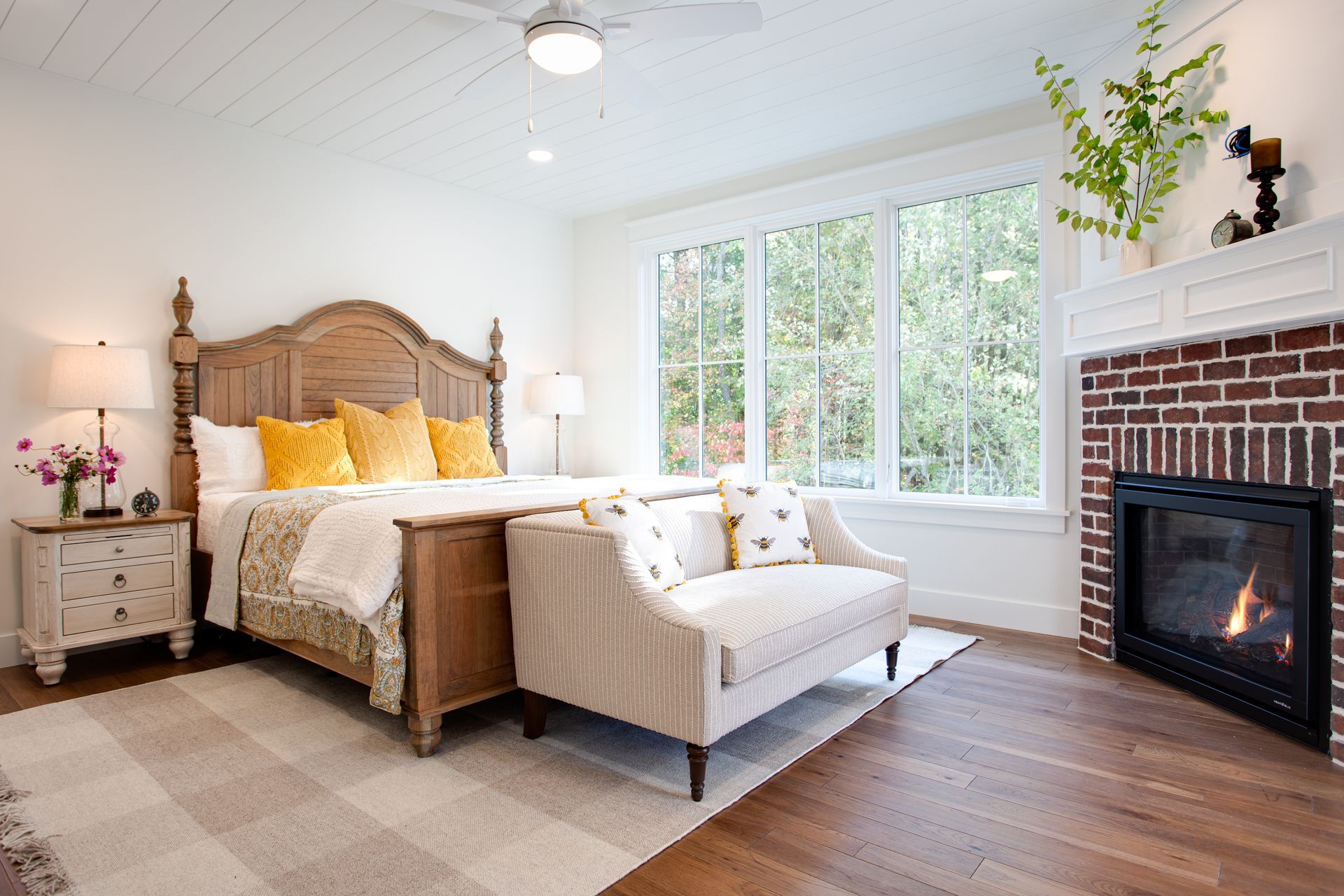 Bright bedroom with a wooden bed, beige chair, large window, and brick fireplace on a white wall.