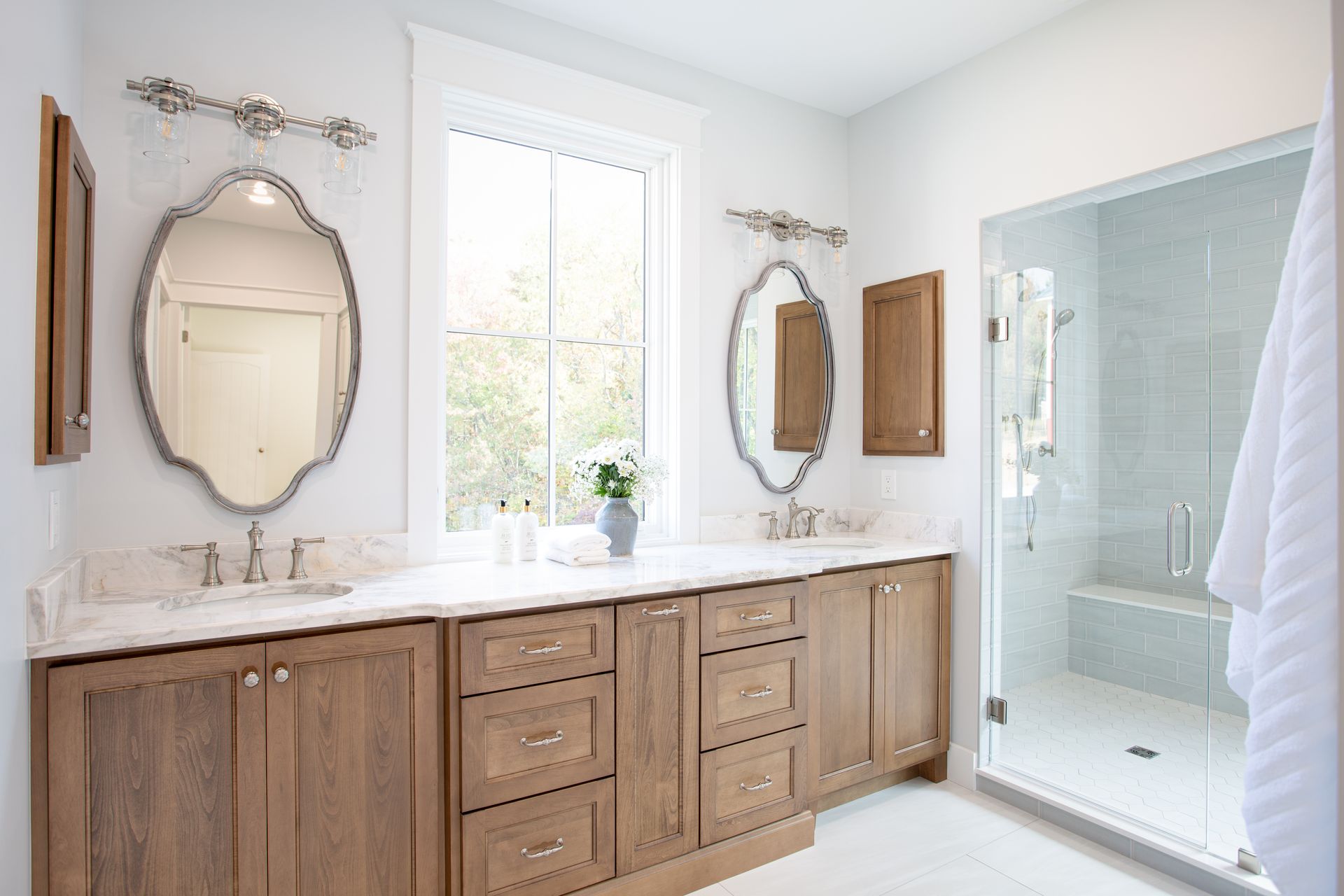 Bright bathroom with wood vanity, marble countertop, oval mirrors, and glass shower enclosure.