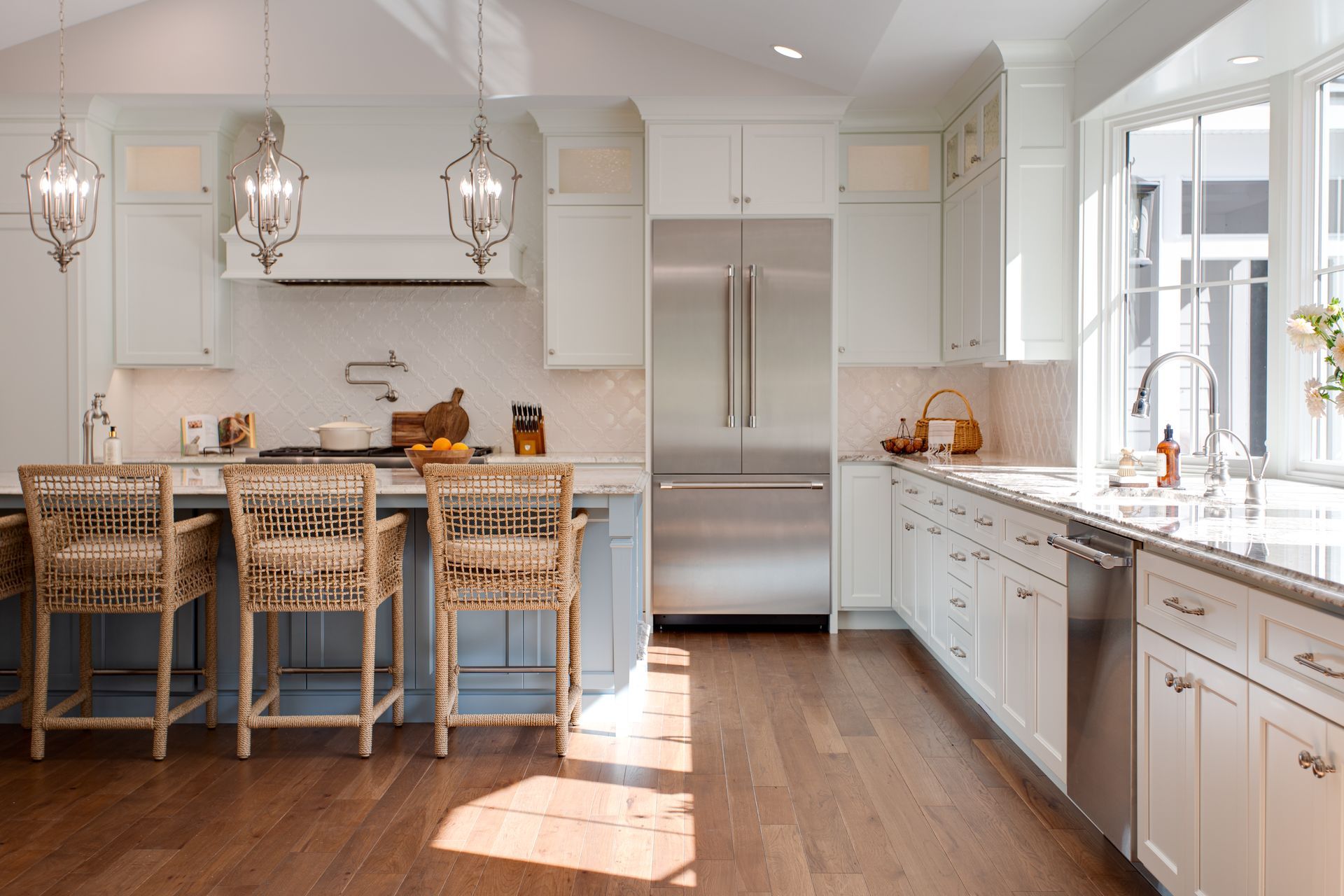 Bright modern kitchen with white cabinets, island stools, and stainless steel refrigerator, lit by large windows.