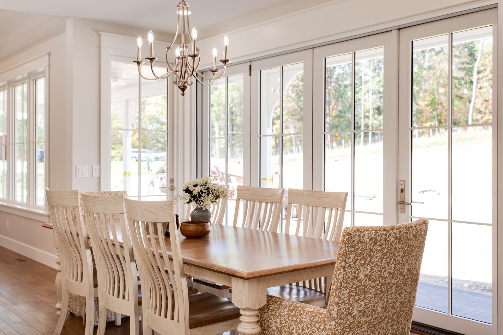 Bright dining room with a long wooden table, chandelier, white chairs, and large glass doors opening outside.