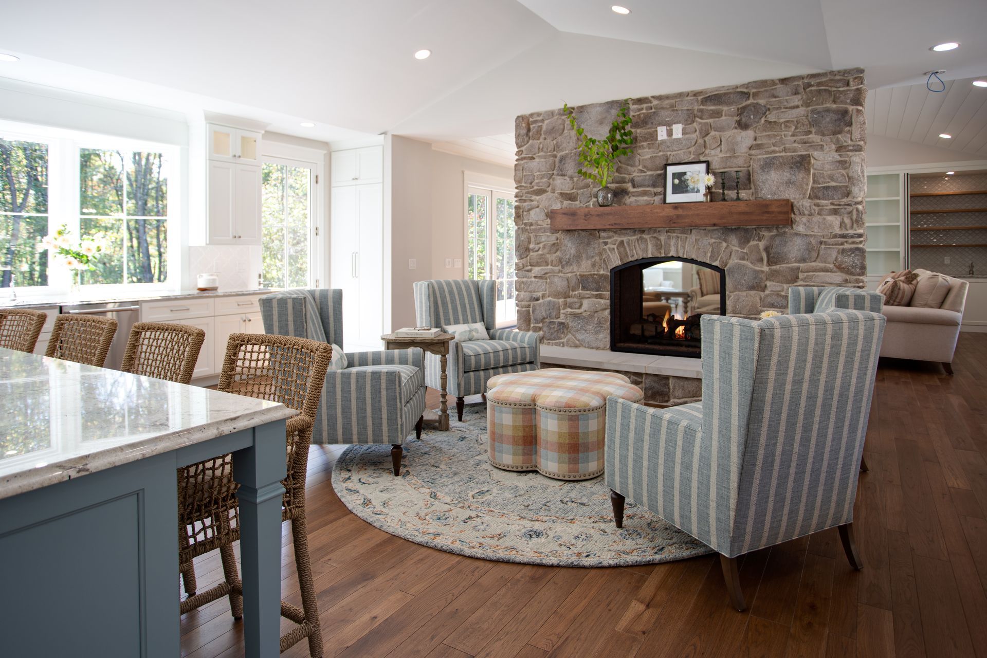Bright living room with stone fireplace, plaid armchairs, round rug, and large windows.