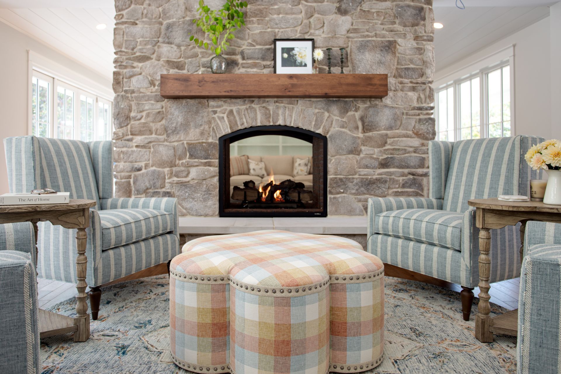 Cozy stone fireplace seating area with striped armchairs and plaid ottoman on a patterned rug.