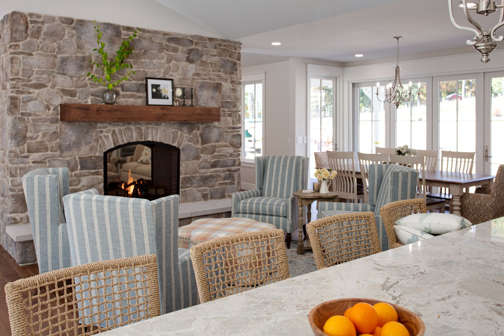 Cozy living room with stone fireplace, armchairs, large windows, and a kitchen island with fruit bowl.