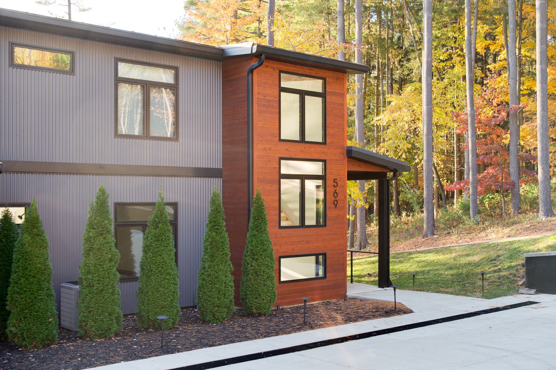 Modern custom home exterior with gray siding, red brick accents, and tall evergreens in autumn.