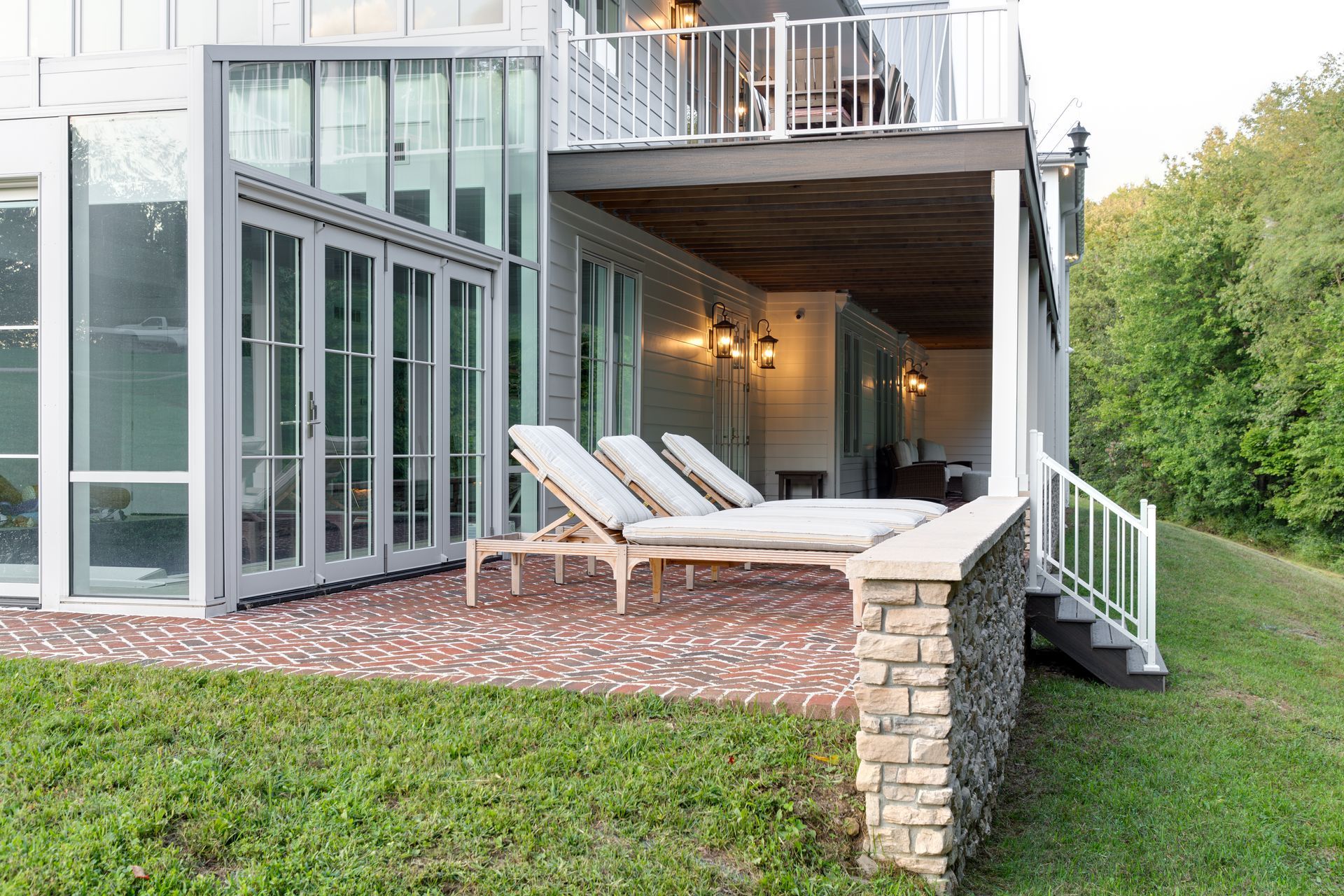 Modern home patio with lounge chairs, brick floor, and glass doors and windows opening to a green yard.