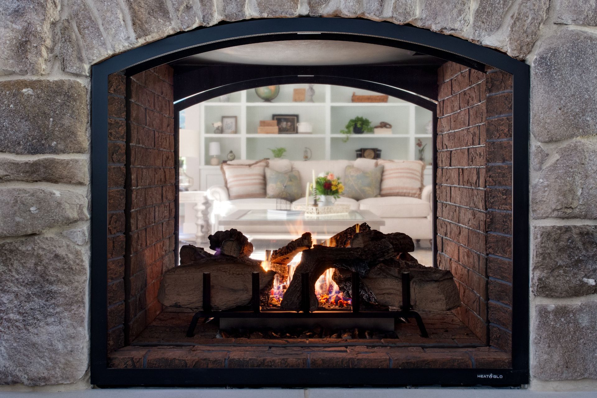 Stone fireplace with a small fire burning in front of a cozy room with a sofa and shelves beyond.