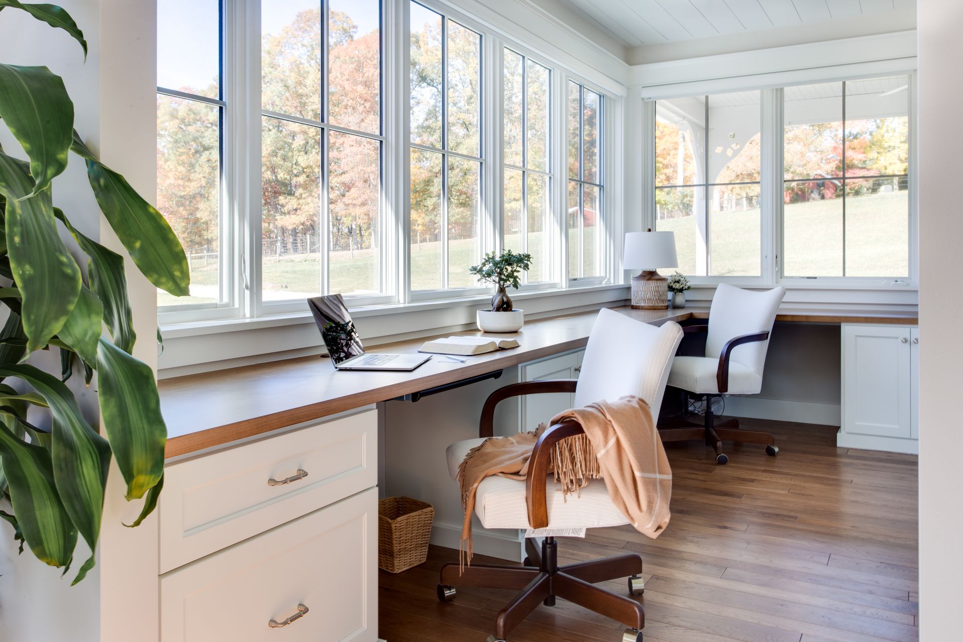 Bright sunroom home office with a long desk, rolling chair, laptop, and large windows.