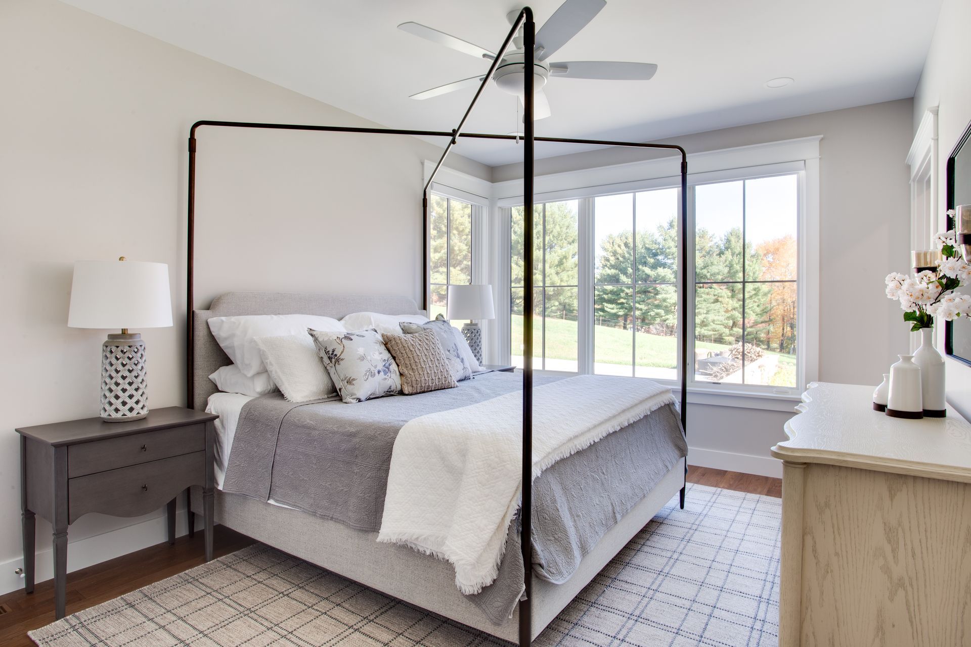 Bright bedroom with canopy bed, gray bedding, bedside table, and large windows overlooking greenery.