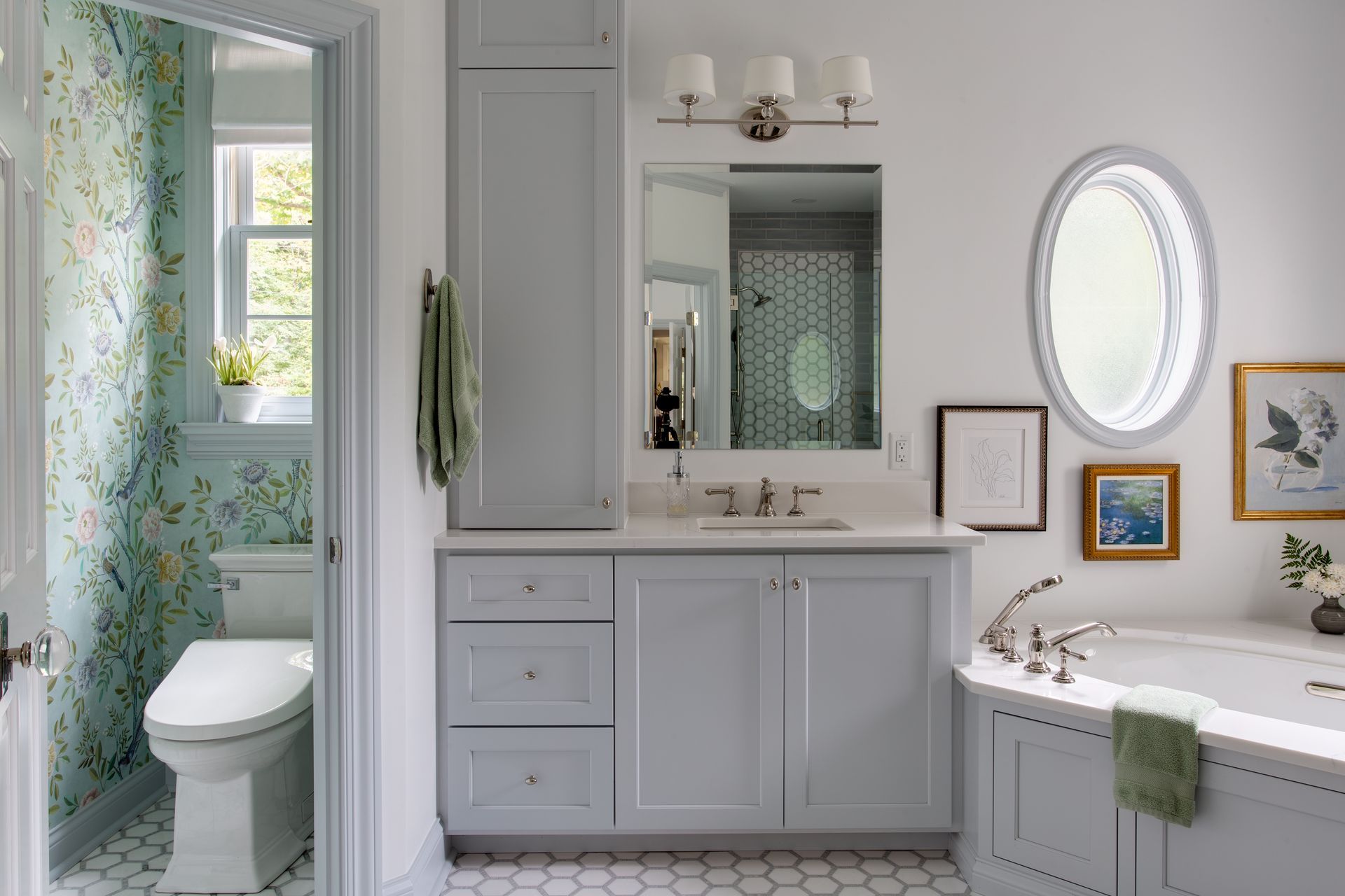 Bright bathroom with white vanity, oval mirror, floral shower curtain, and window by the toilet.