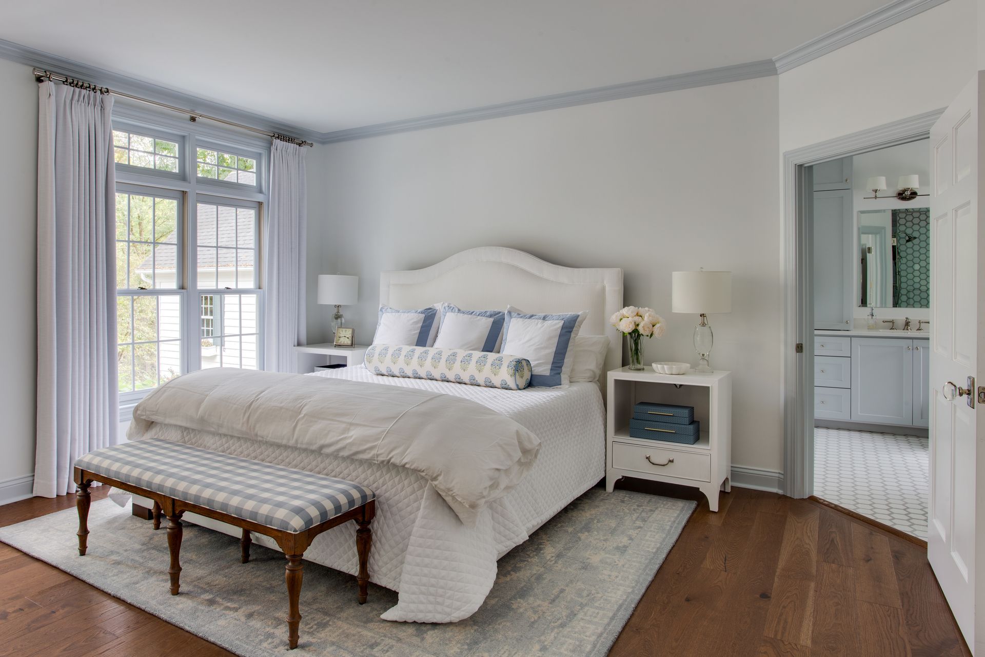 Bright bedroom with a white bed, bench, bedside tables, and large windows by an adjoining bathroom.