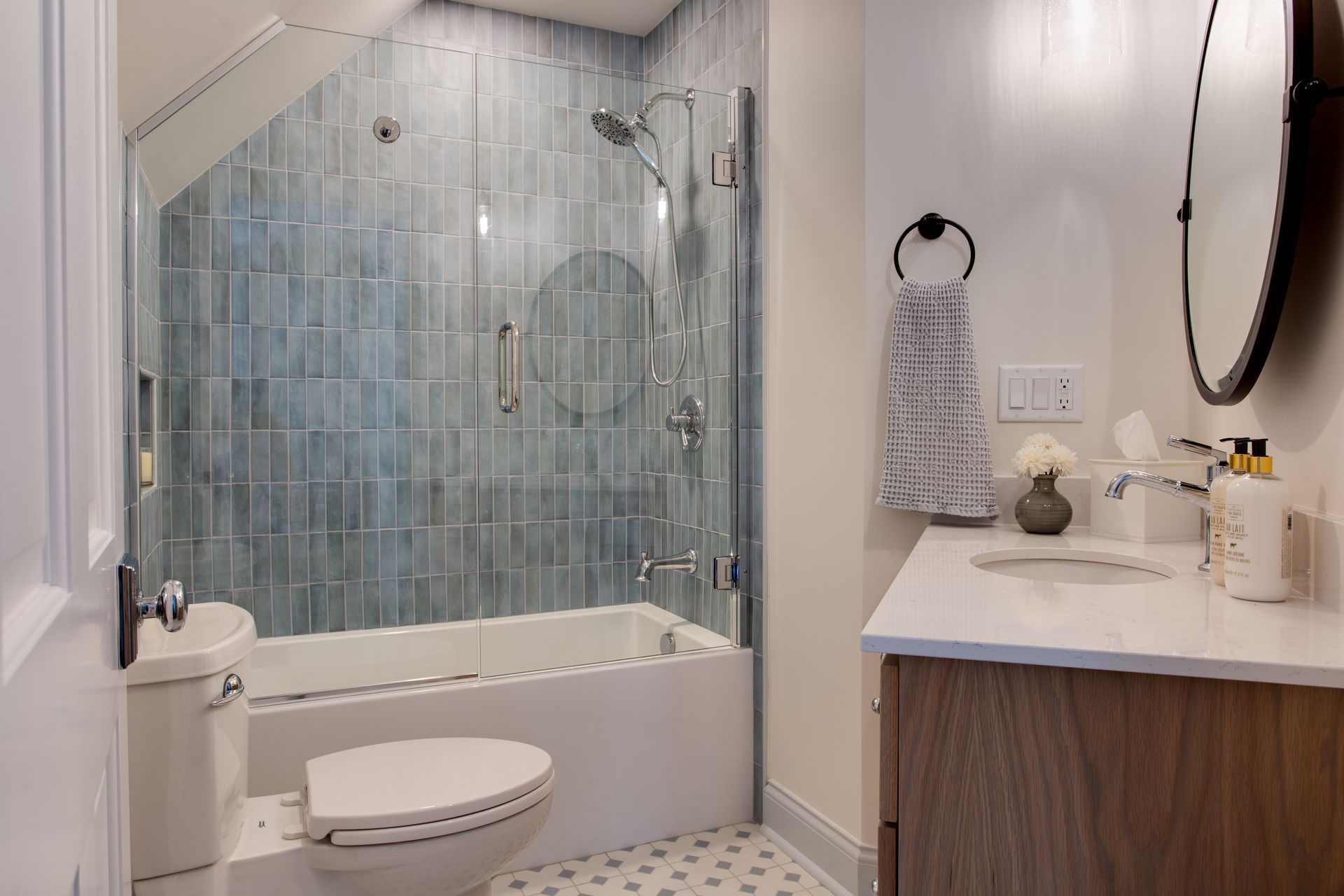 Bathroom with glass-enclosed tub-shower, blue tile, and white vanity with round mirror.