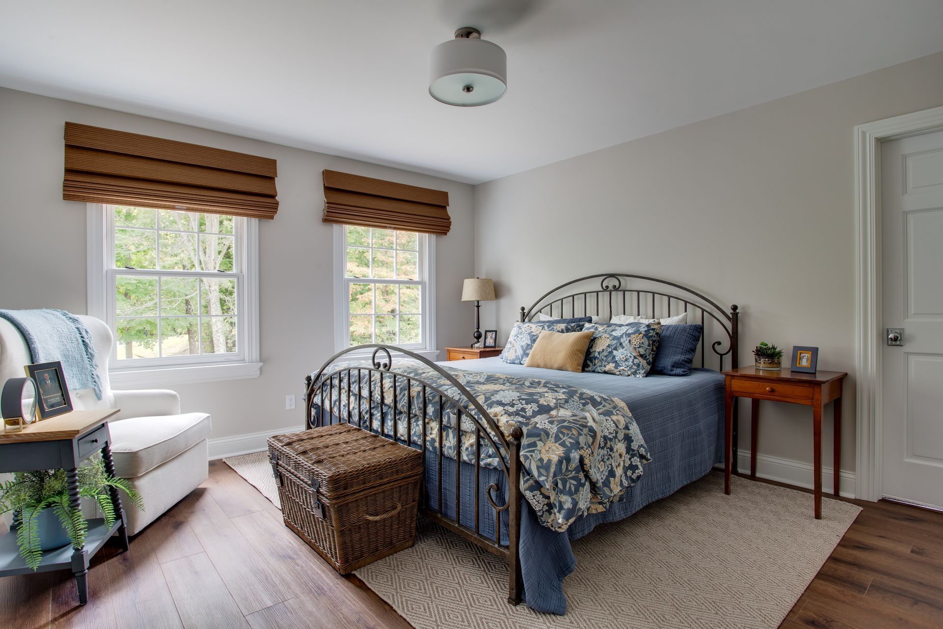 Bright bedroom with black metal bed, blue bedding, wicker ottoman, two windows, and a wooden side table.