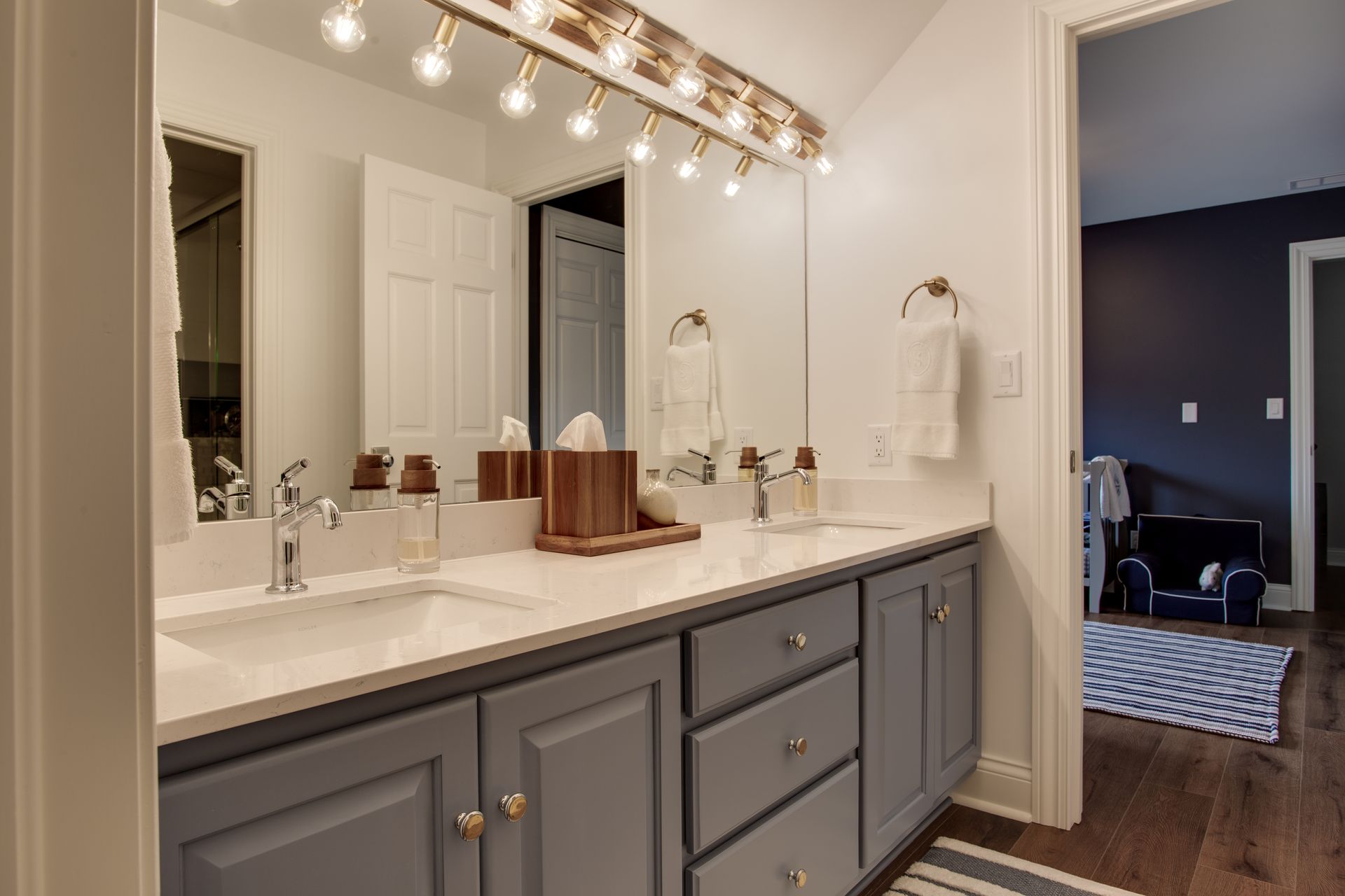Modern bathroom vanity with gray cabinets, large mirror, bright lights, and an open doorway to a bedroom.