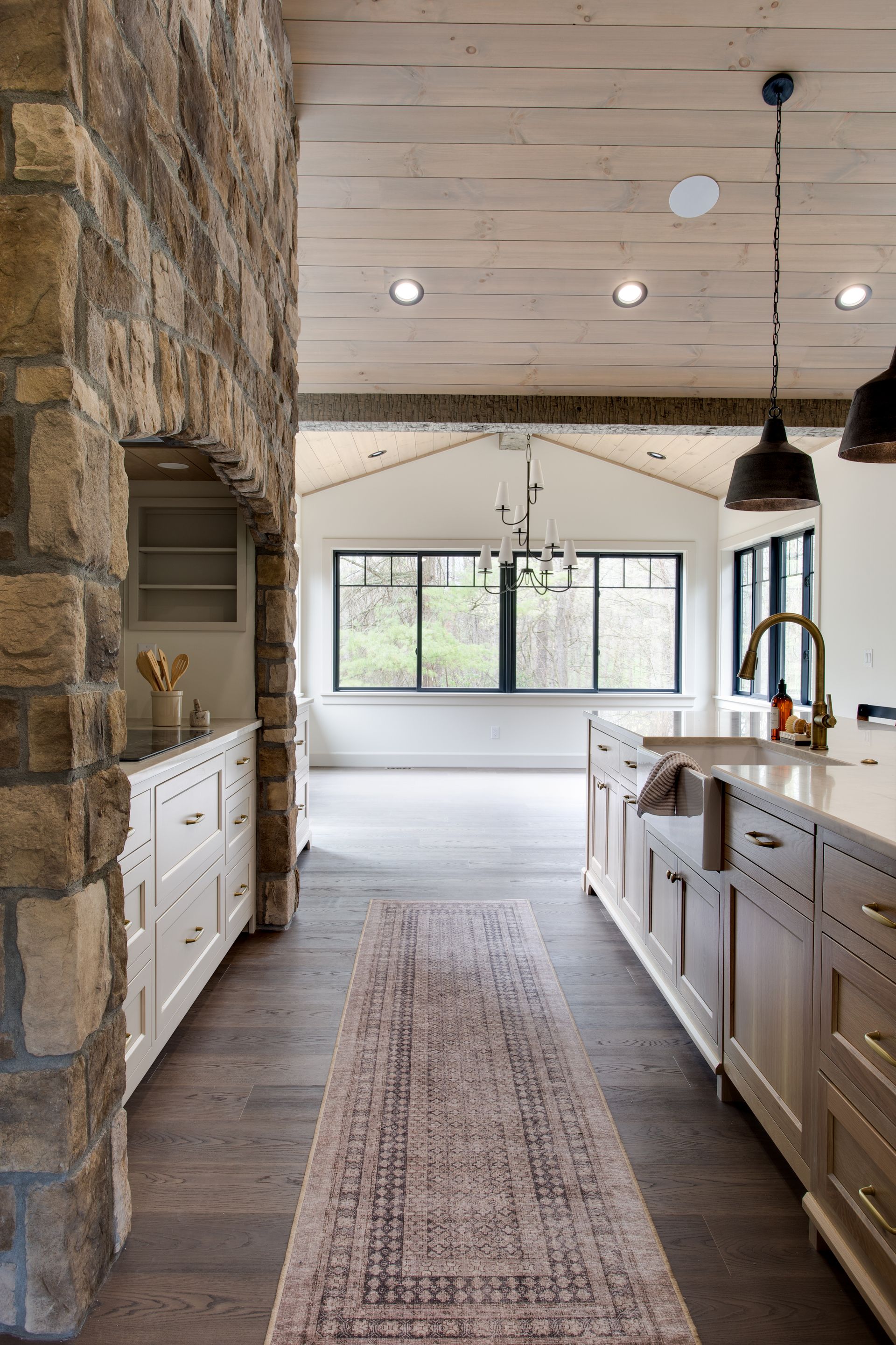 Rustic kitchen with stone wall, white cabinets, island, and large windows in a bright open room.