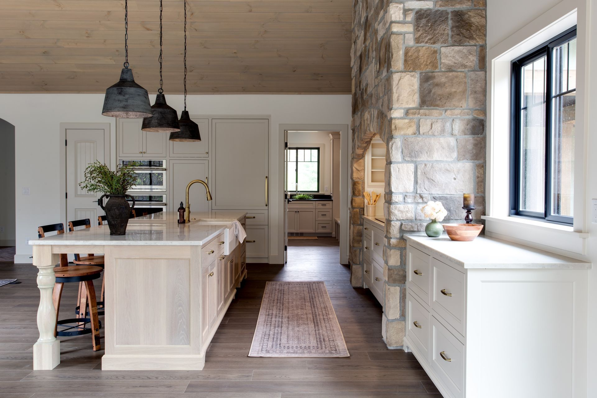 Bright modern kitchen with white island, pendant lights, stone wall, and windowed sideboard.