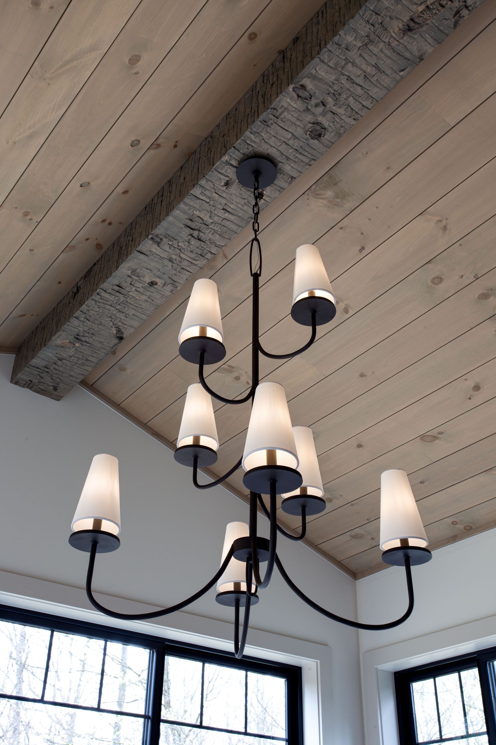 Black chandelier attached to reclaimed wood beam with six white shades hanging from a vaulted wood ceiling above windows.