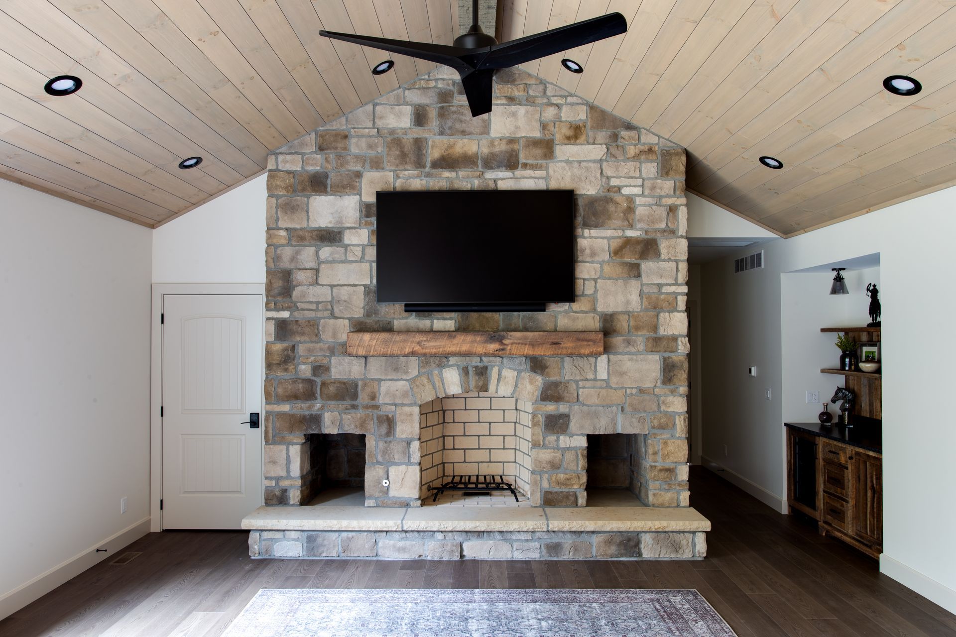 Living room of a luxury home with vaulted ceiling, stone fireplace wall, mounted TV, and white built-in cabinets.