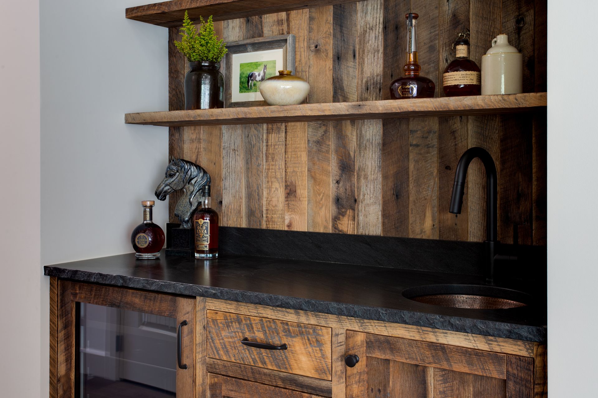 Rustic kitchen bar with dark countertop, wood shelves, sink, and decorative bottles and plants.