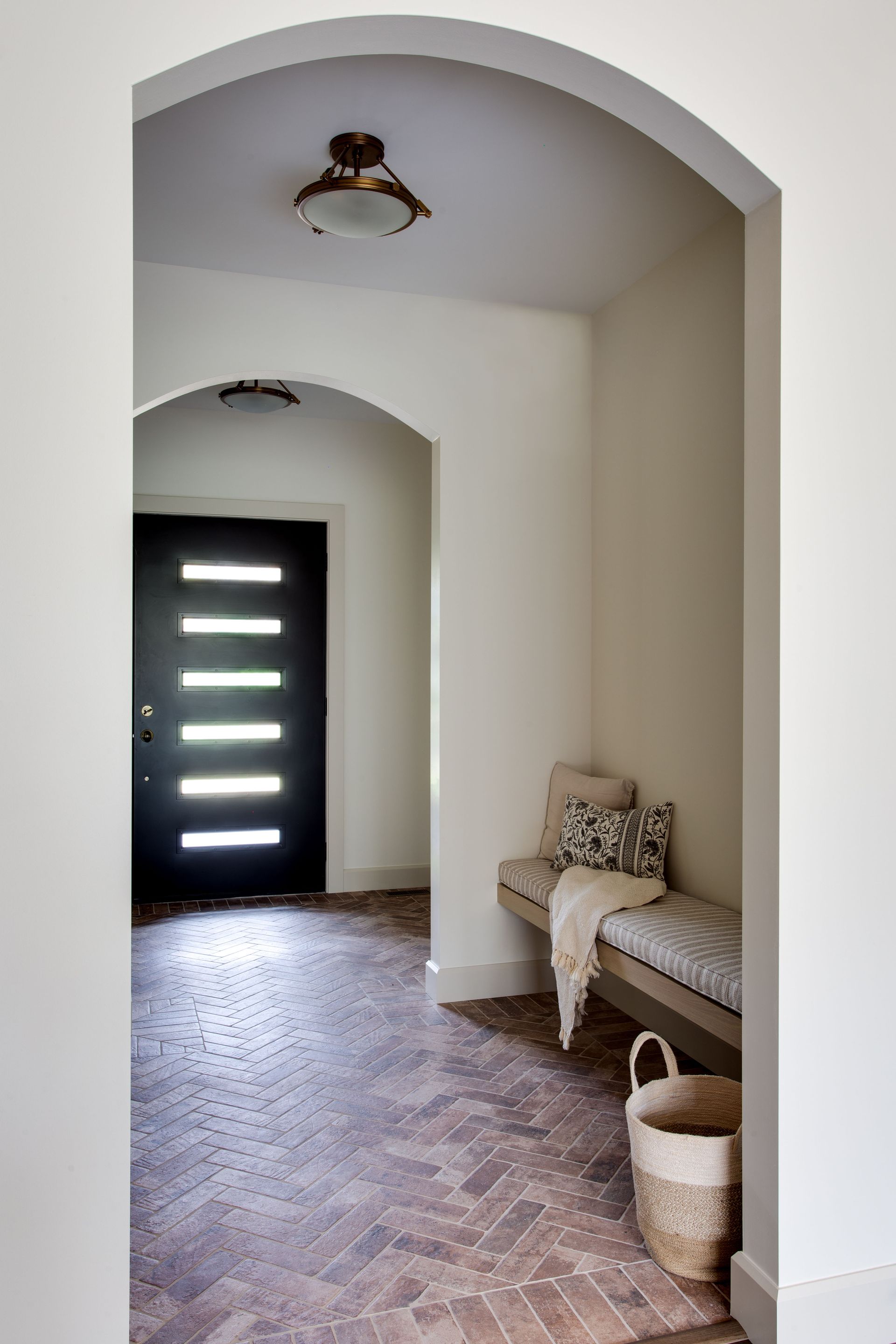 Arched hallway with bench, basket, and black door in a bright, tiled entryway.