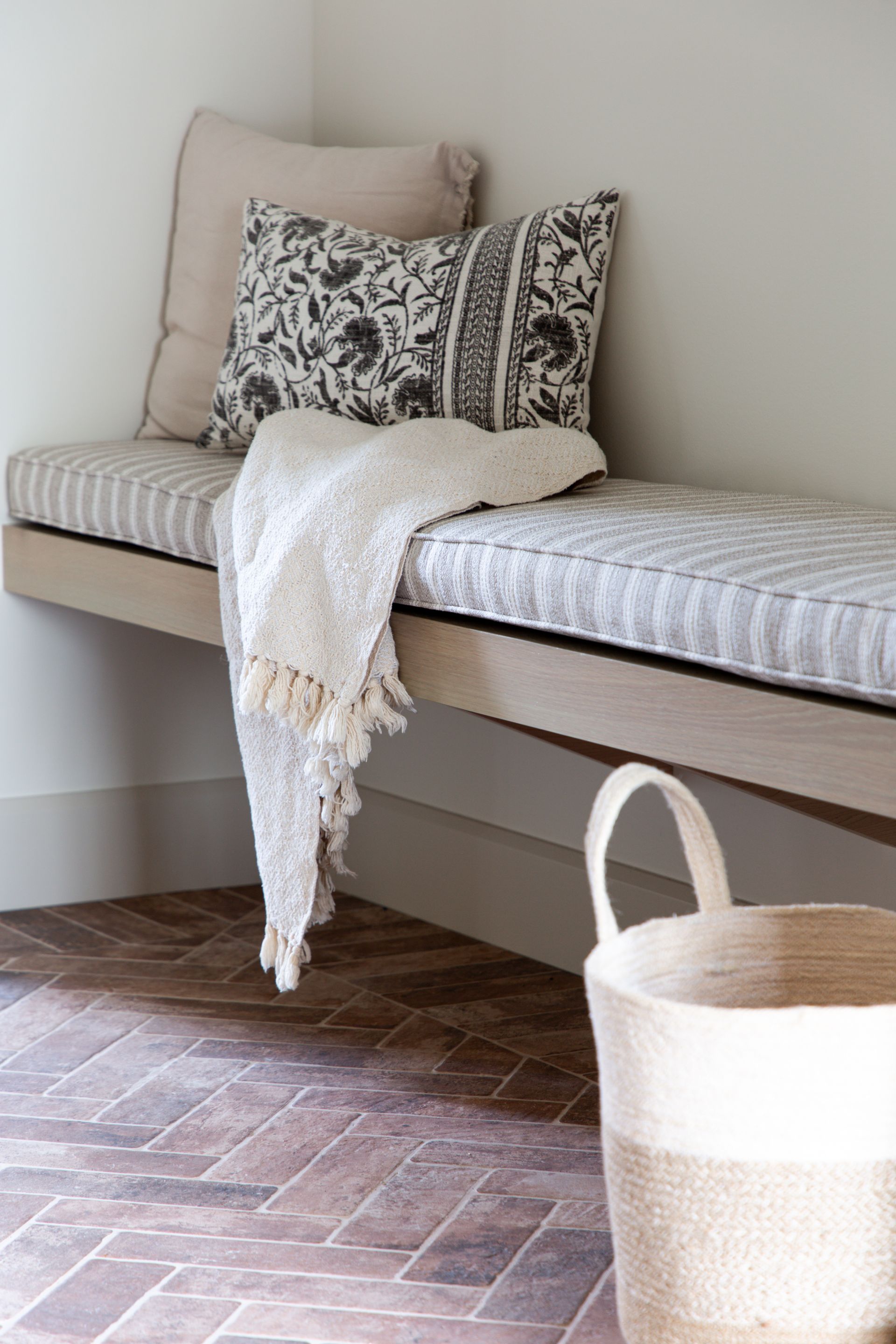 Striped bench with cushion, patterned pillows, throw blanket, and woven basket on a brick floor.