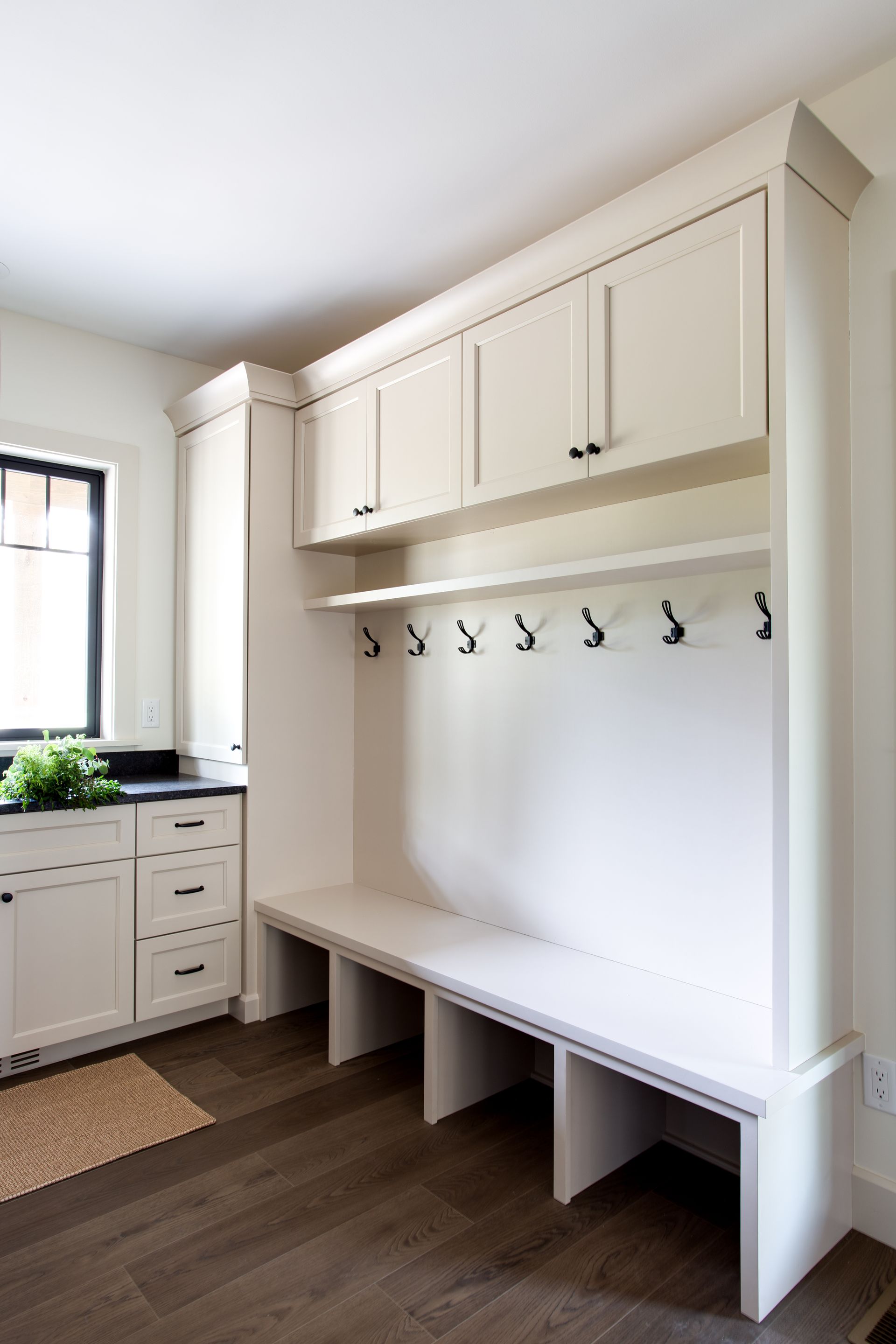 Built-in mudroom bench with white cabinets and coat hooks beside a window and sink.
