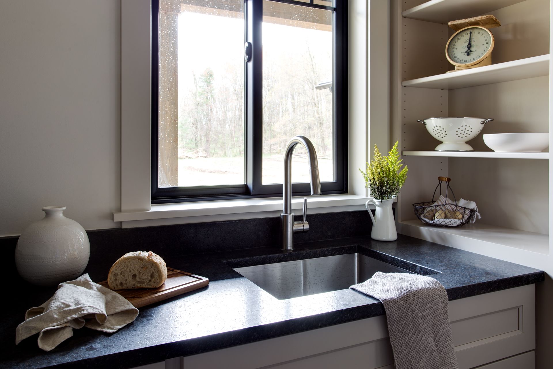 Modern kitchen sink by a window with black countertop, faucet, shelves, and decorative items.