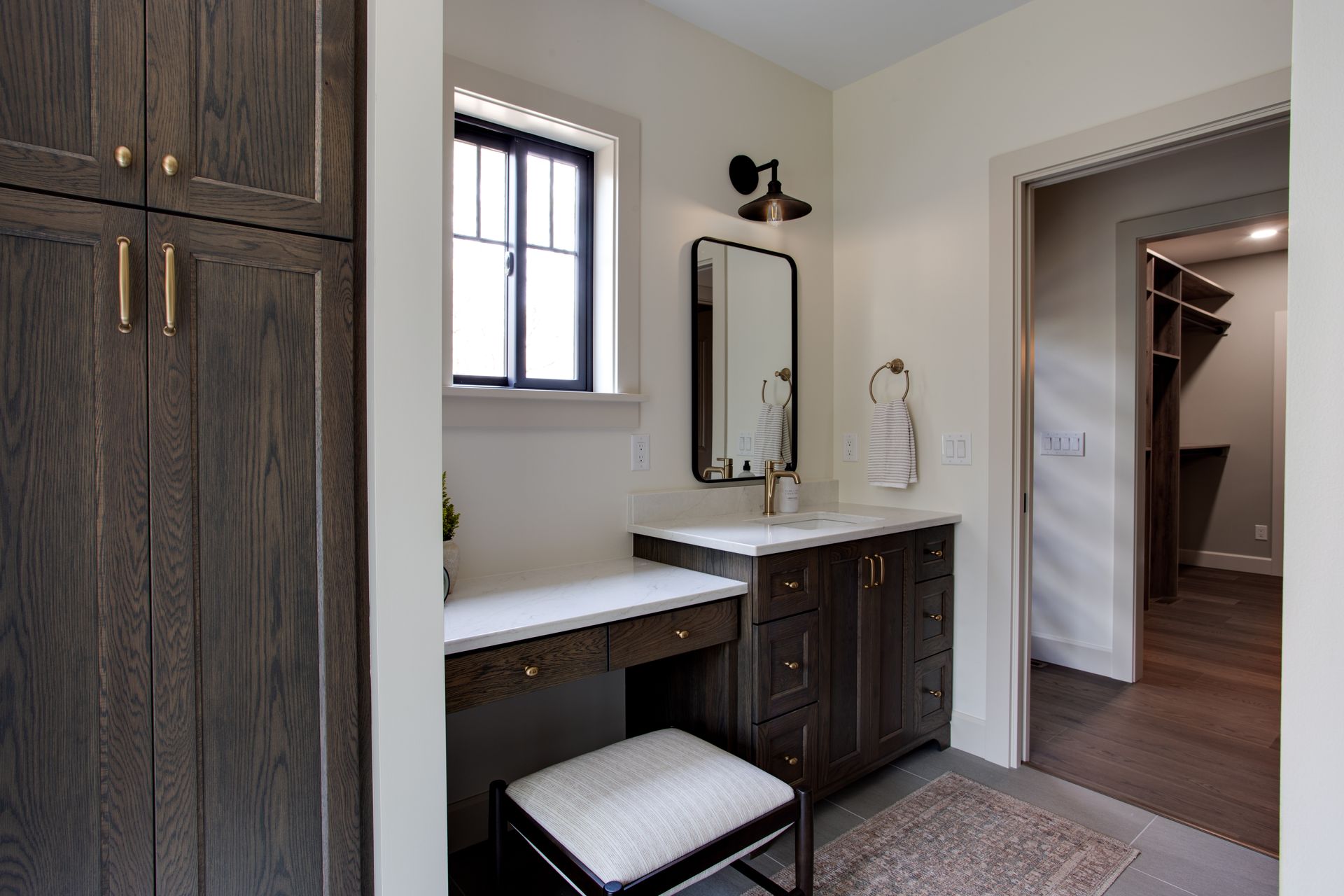 Bathroom vanity with dark wood cabinets, mirror, window, and upholstered stool in a beige room
