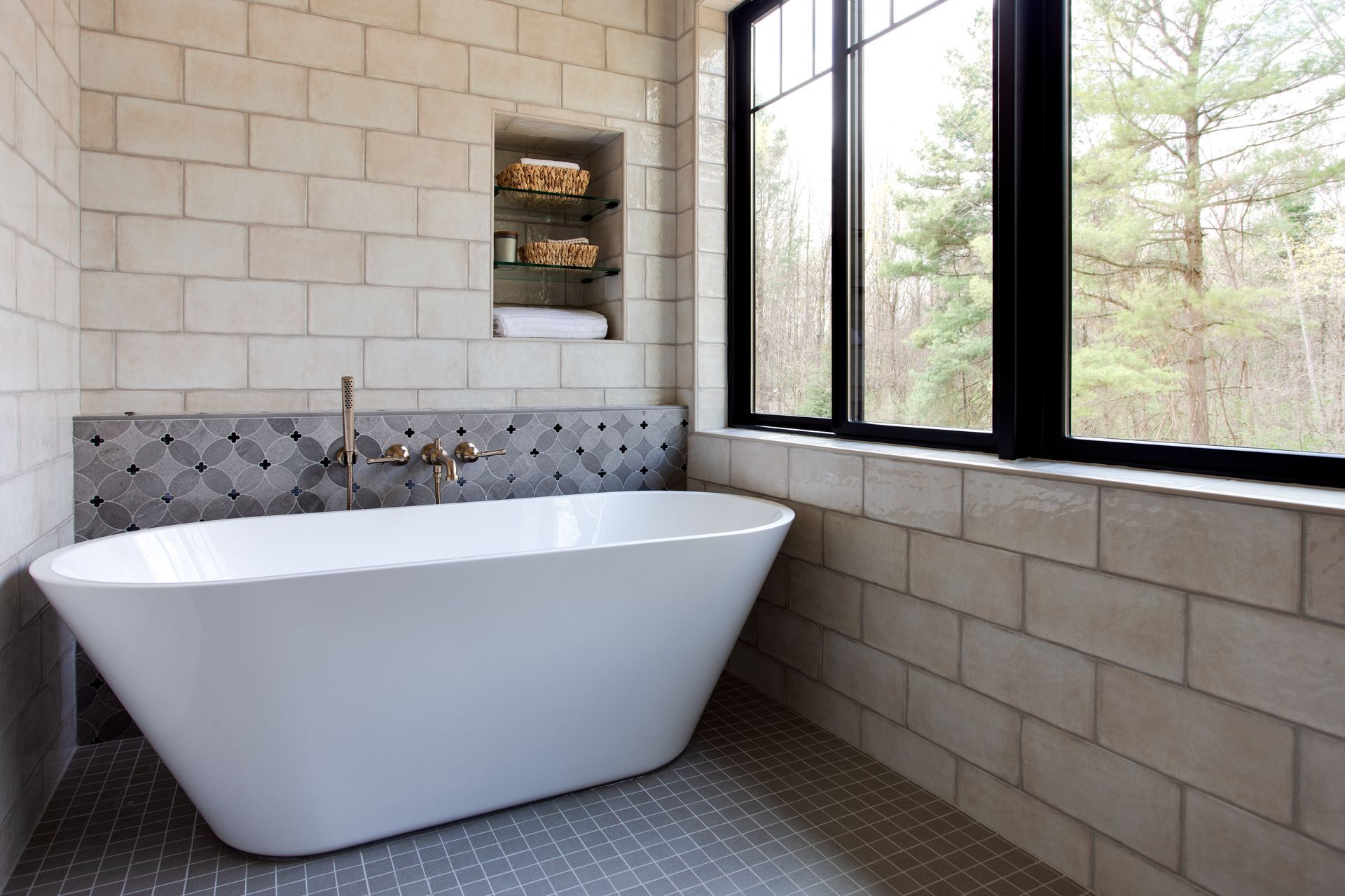 Modern bathroom with white freestanding tub, tiled walls, and large windows overlooking trees.