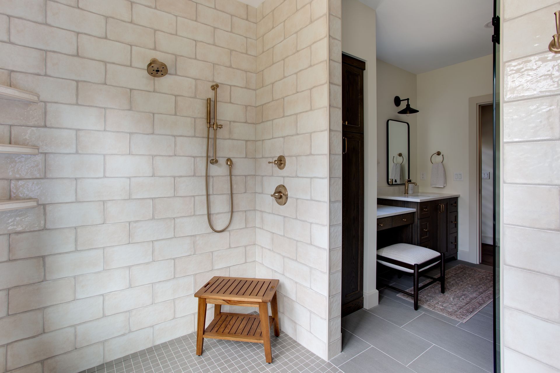 Walk-in shower with white tile, brass fixtures, and a wooden stool beside an open bathroom vanity.