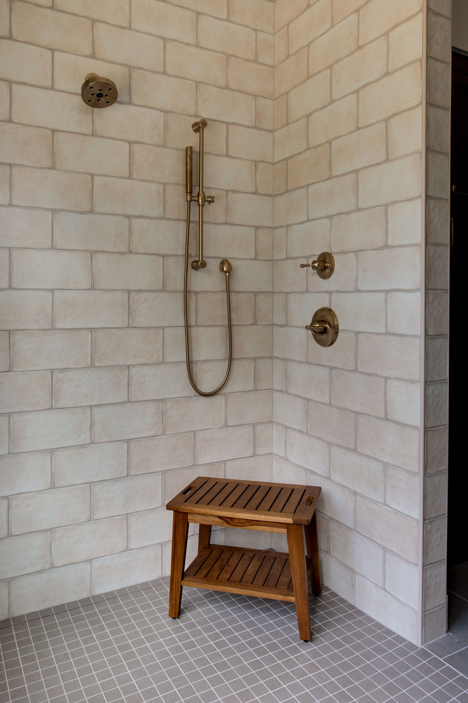 Tile shower corner with brass fixtures, handheld shower, and wooden stool on a pebble floor.