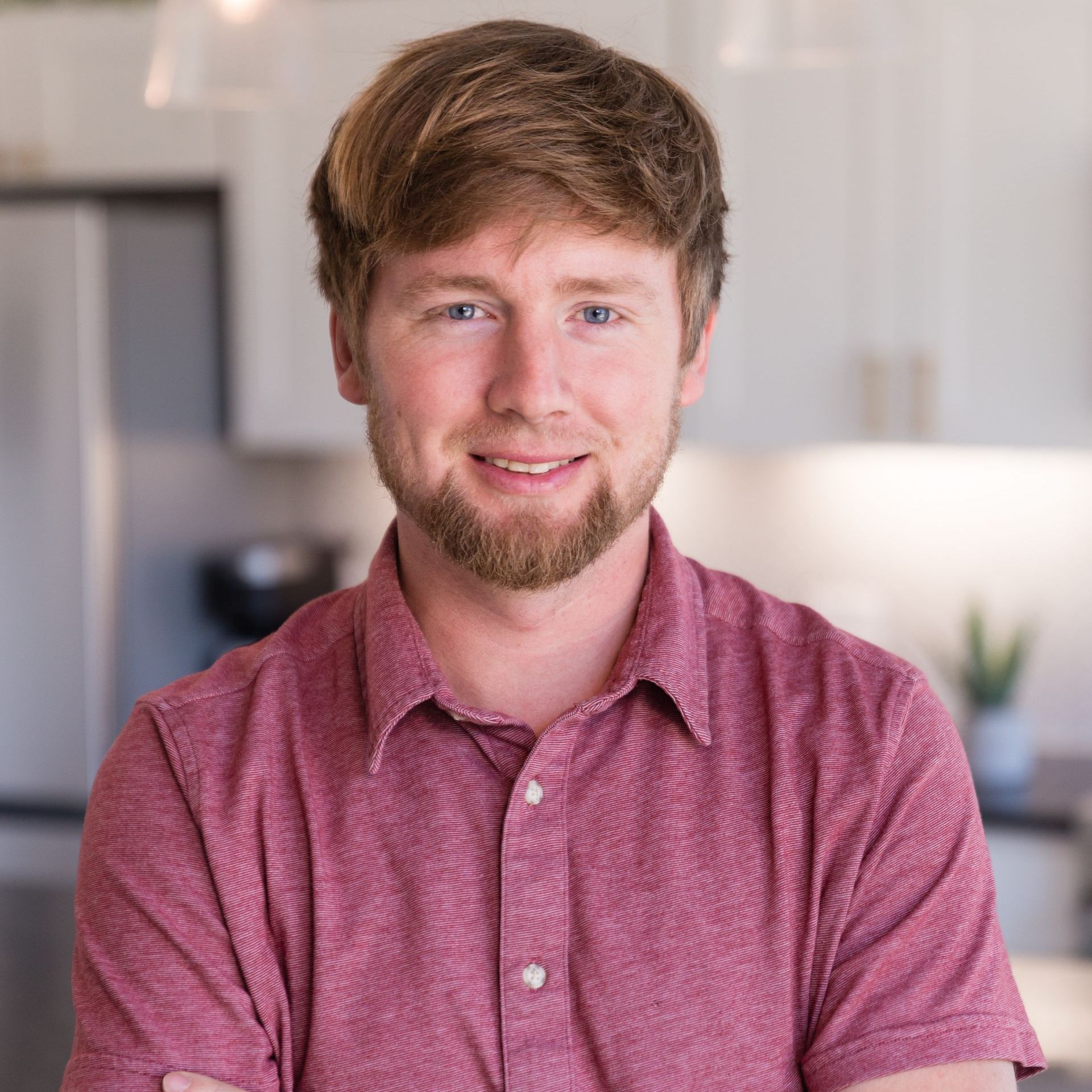 A man with a beard wearing a red shirt is standing in a kitchen with his arms crossed.