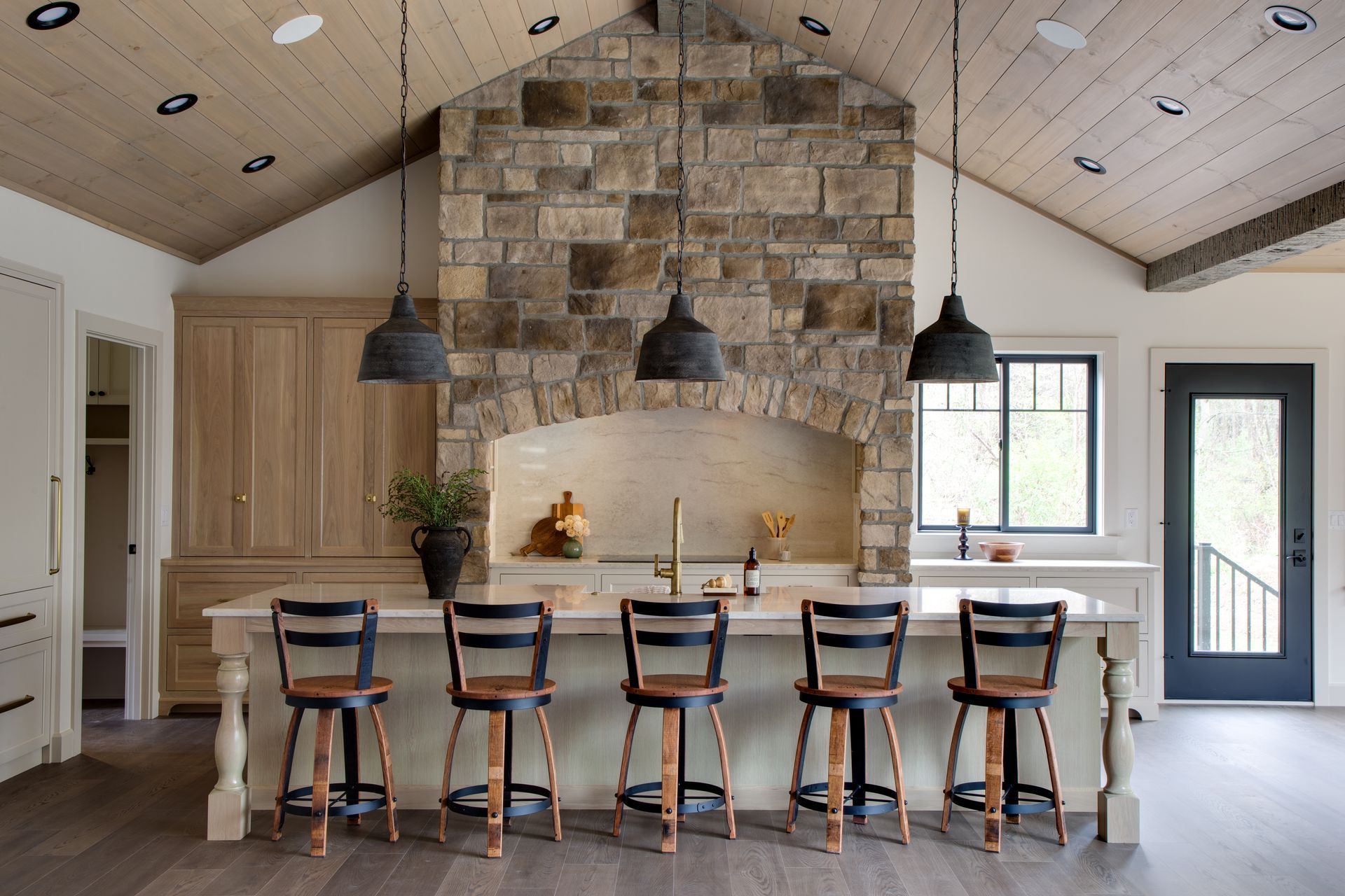 Kitchen with stone range hood, island with stools, wood ceiling, and black pendant lights.