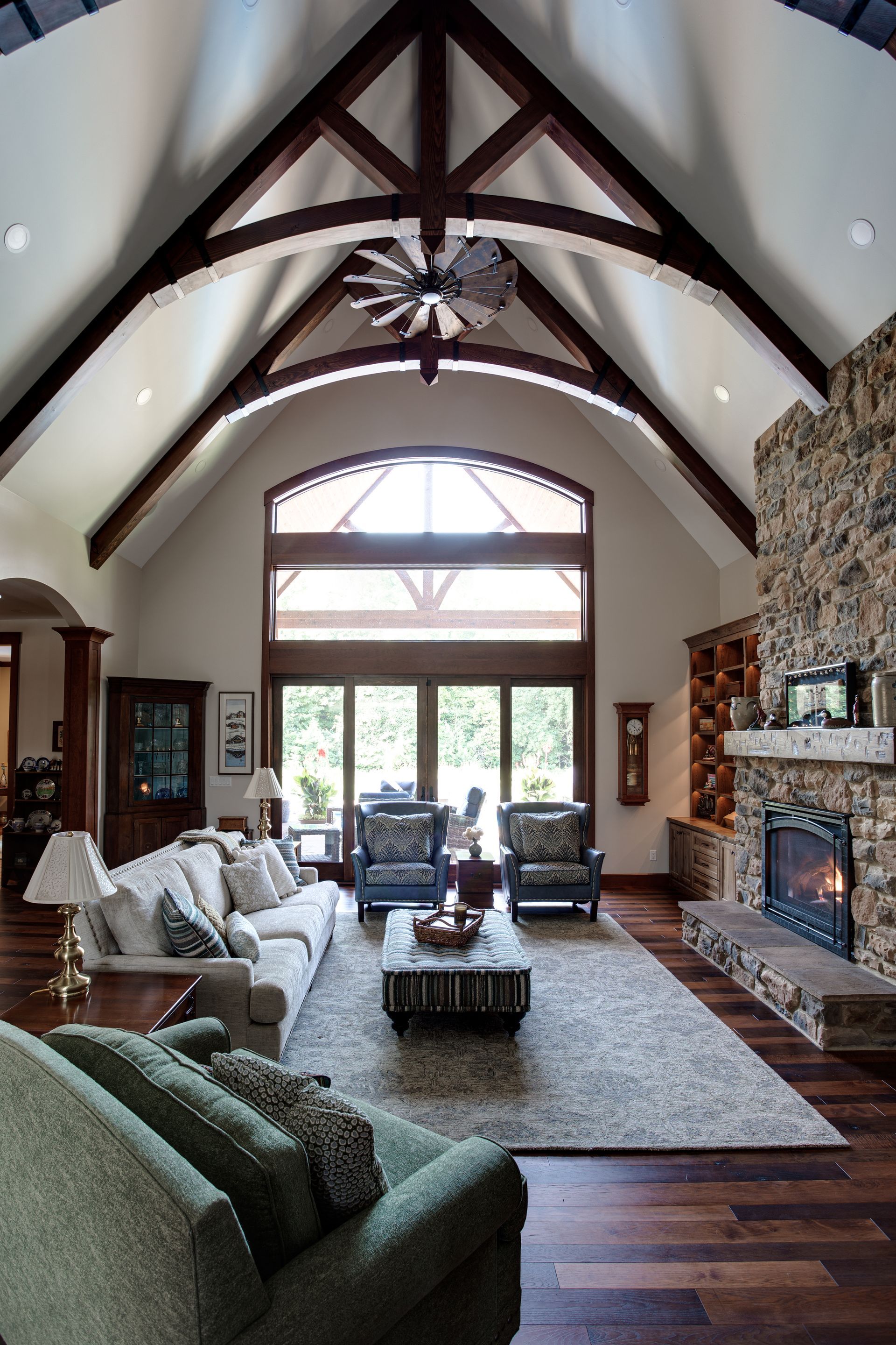 Spacious living room with vaulted wood-beam ceiling, stone fireplace, and large windows overlooking trees
