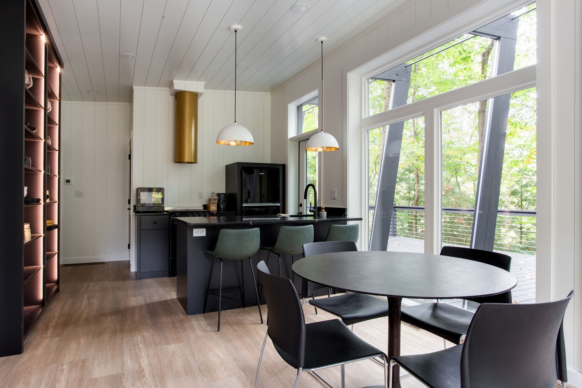 Modern open-plan kitchen and dining area with black furnishings, wood floors, and large windows overlooking trees