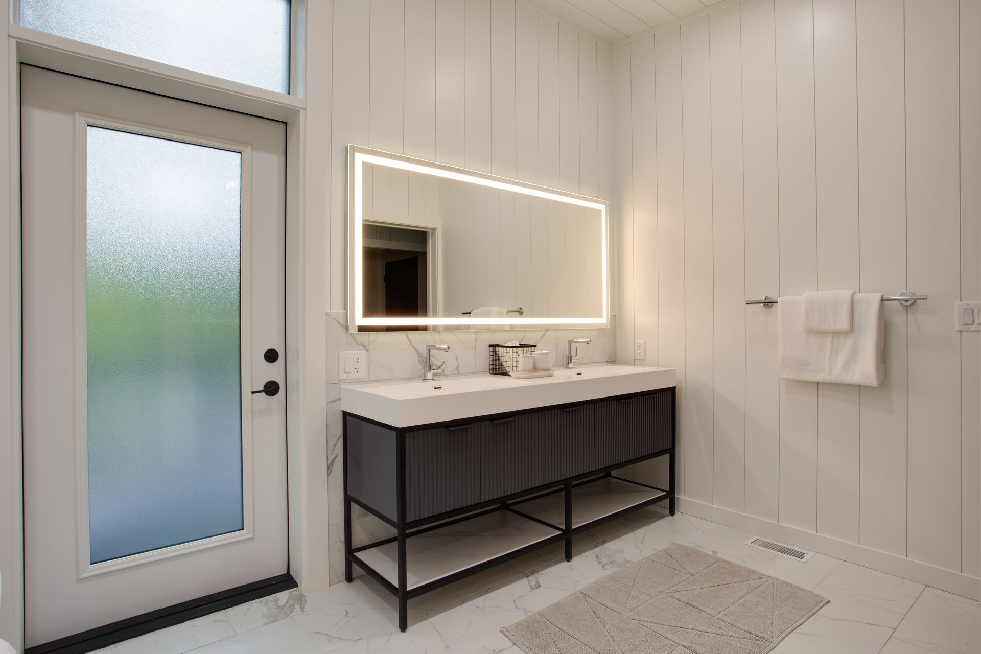 Modern bathroom with illuminated mirror, dark vanity, frosted glass door, and white tiled walls