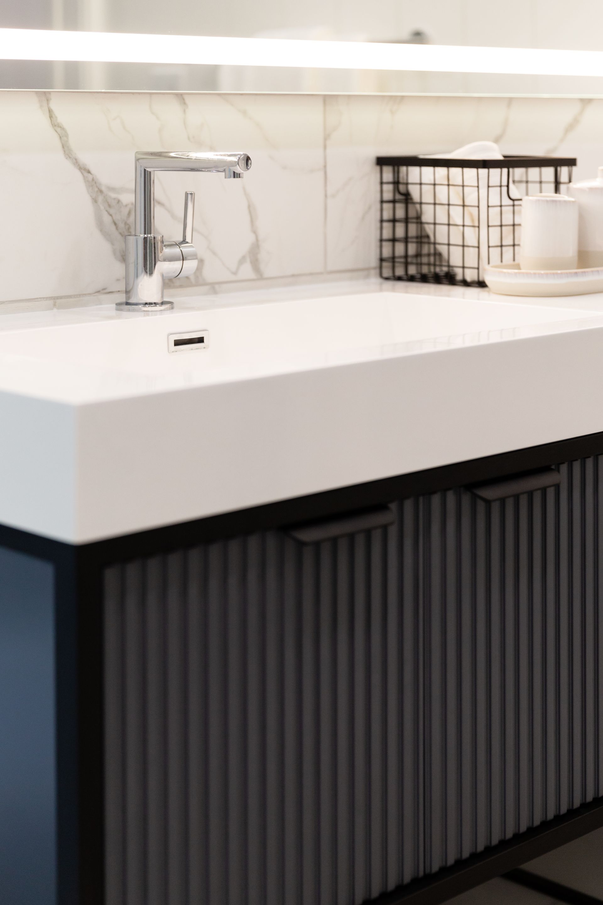 Modern bathroom vanity with white countertop, chrome faucet, black ribbed cabinet, and wire basket decor