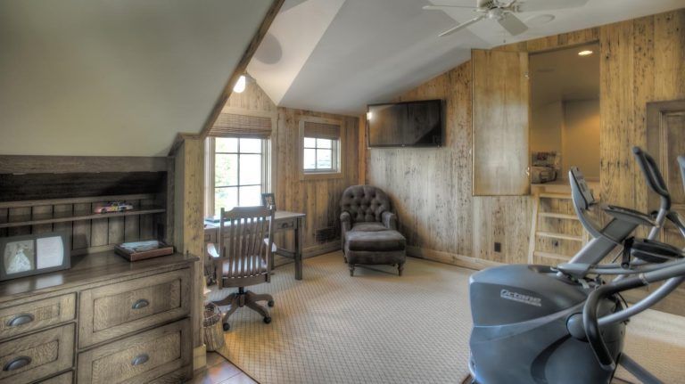 A room with a desk , chair , ottoman , exercise bike and television.