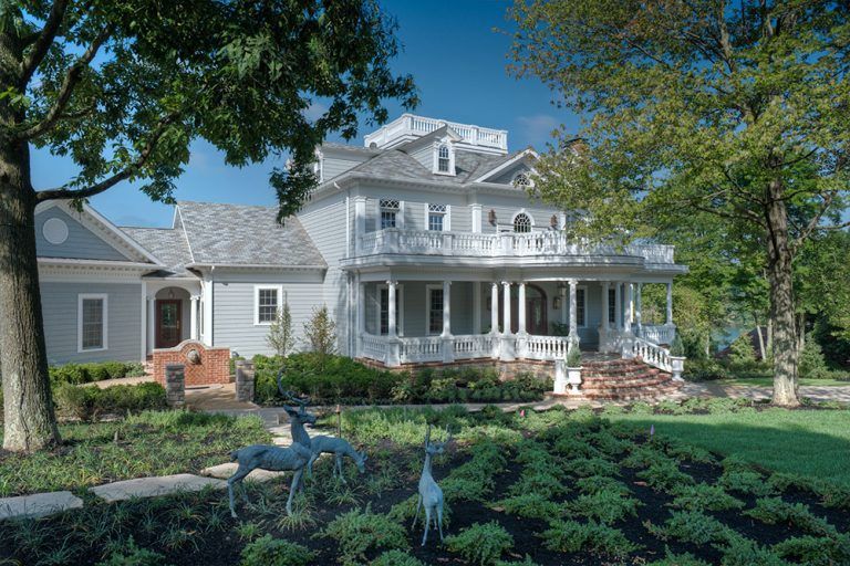 A large white house with a large porch is surrounded by trees and bushes.