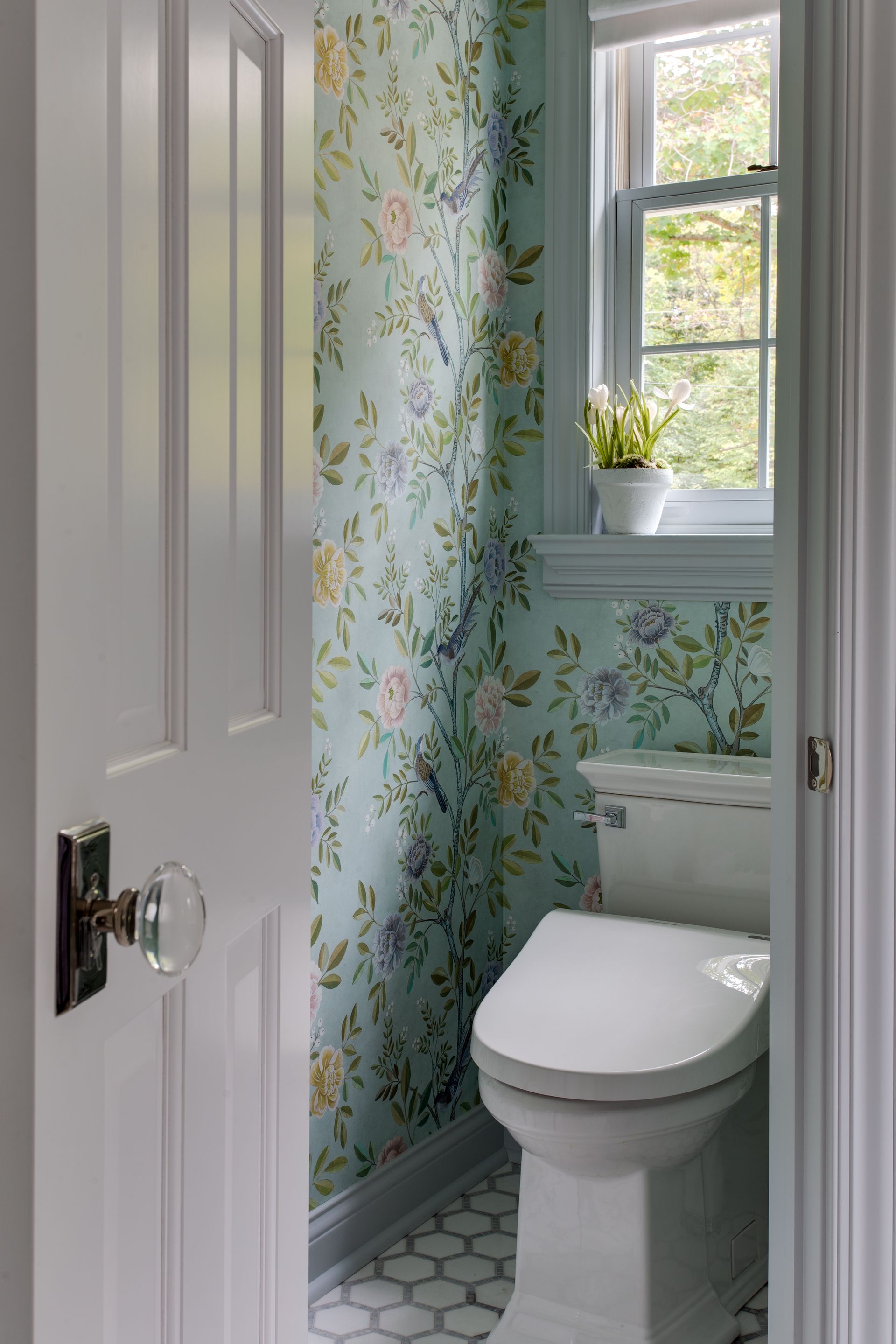 A bathroom with a toilet and a window with floral wallpaper.