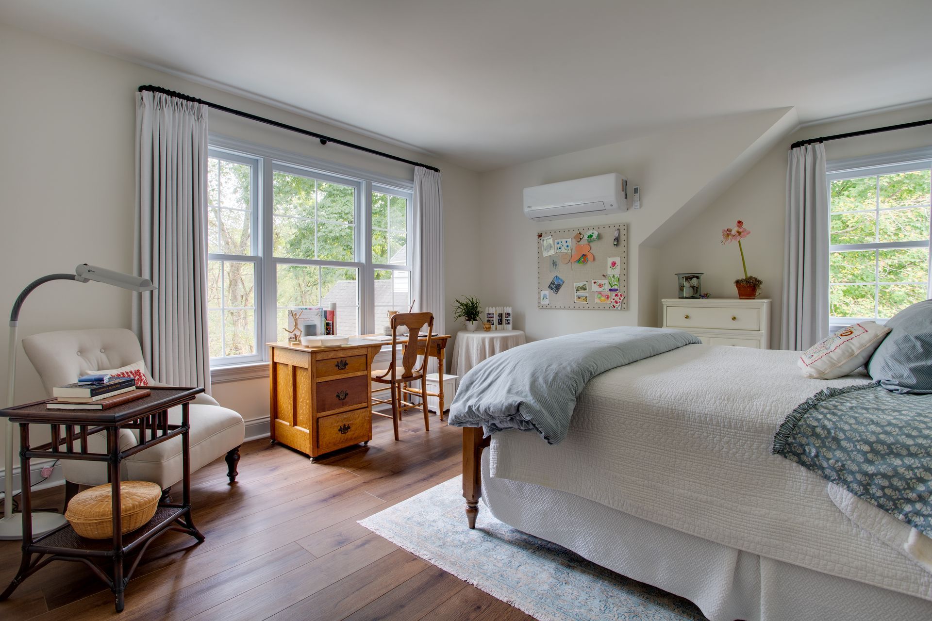 Bright bedroom with twin beds, large windows, wooden dresser, and soft gray-blue decor.
