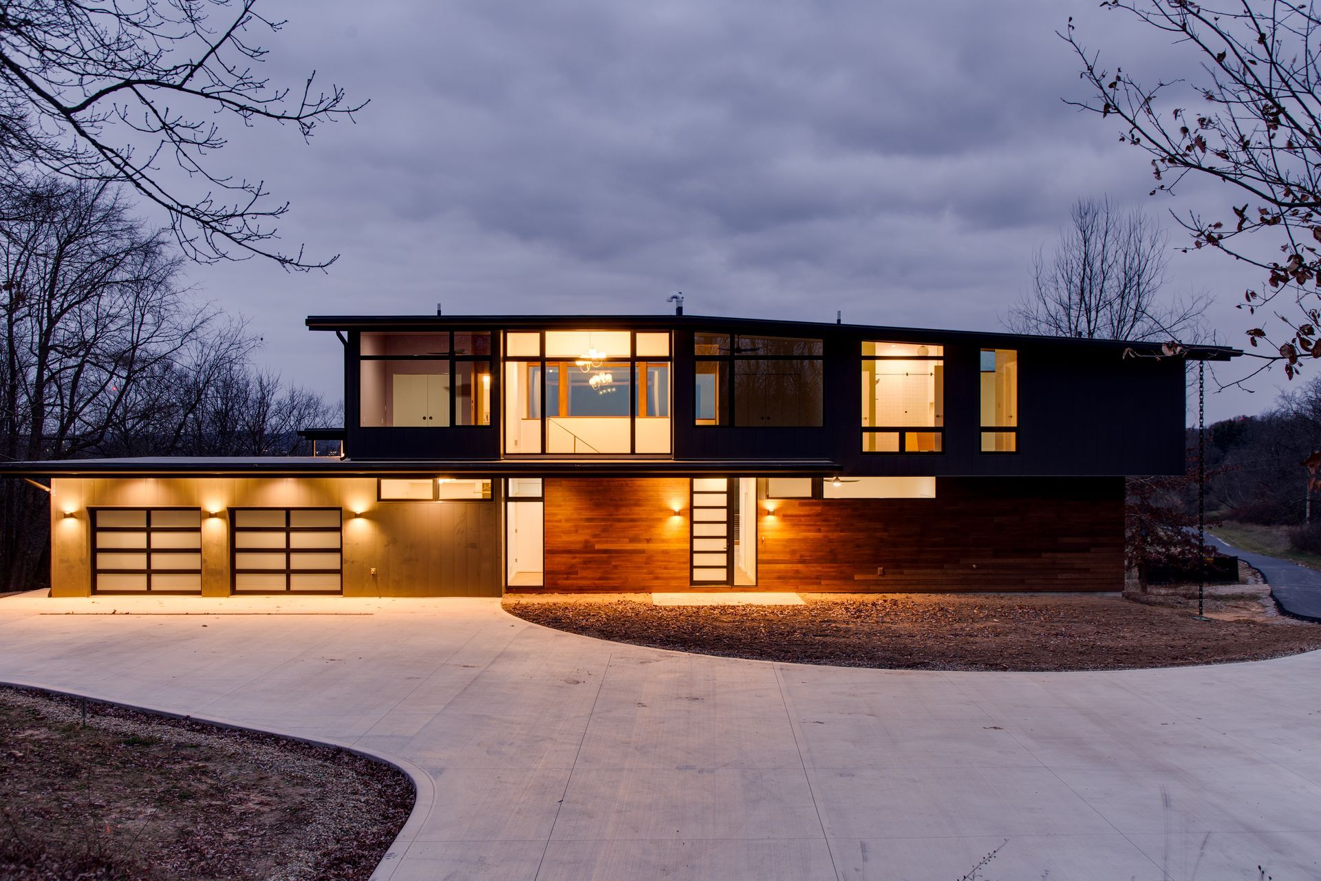Modern two-story house at dusk with warm interior lights and a large lit driveway